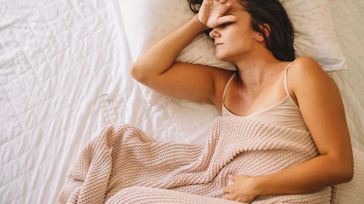 Eine junge Frau liegt krank im Bett. Da sie mit einem Arm ihre Stirn hält und mit dem anderen ihr Unterleib, sieht es danach aus, als habe sie dort in beiden Bereichen Schmerzen, sodass sie vermutlich an menstrueller Migräne leidet.