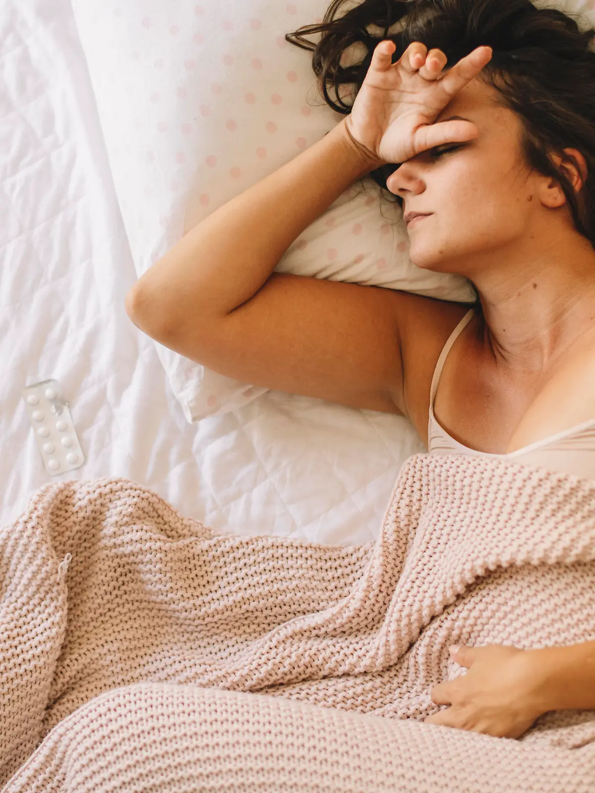 Eine junge Frau liegt krank im Bett. Da sie mit einem Arm ihre Stirn hält und mit dem anderen ihr Unterleib, sieht es danach aus, als habe sie dort in beiden Bereichen Schmerzen, sodass sie vermutlich an menstrueller Migräne leidet.