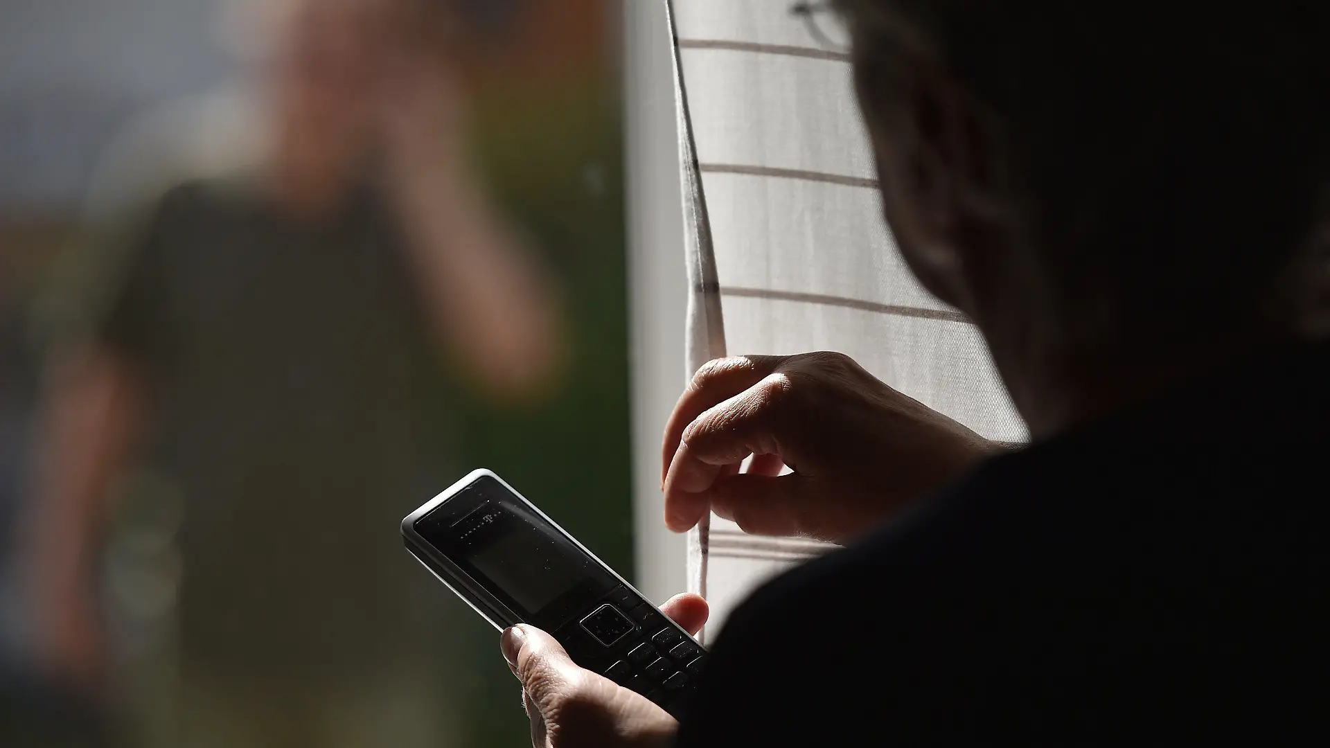 ARCHIV - ILLUSTRATION - 05.07.2016, Bayern, Garmisch-Partenkirchen: Eine Frau steht mit einem Telefon am Fenster, vor dem ein Mann steht (gestellte Szene). Vor zehn Jahren erhitzte in Berlin ein Hilfsangebot für Stalker die Gemüter. Heute ist diese Arbeit im Sinn des Opferschutzes anerkannt. Doch inzwischen suchen mehr Opfer als Täter Rat. (zu "Bilanz - zehn Jahre Hilfsangebote für Stalker in Berlin" vom 03.05.2018) Foto: Angelika Warmuth/dpa +++ dpa-Bildfunk +++