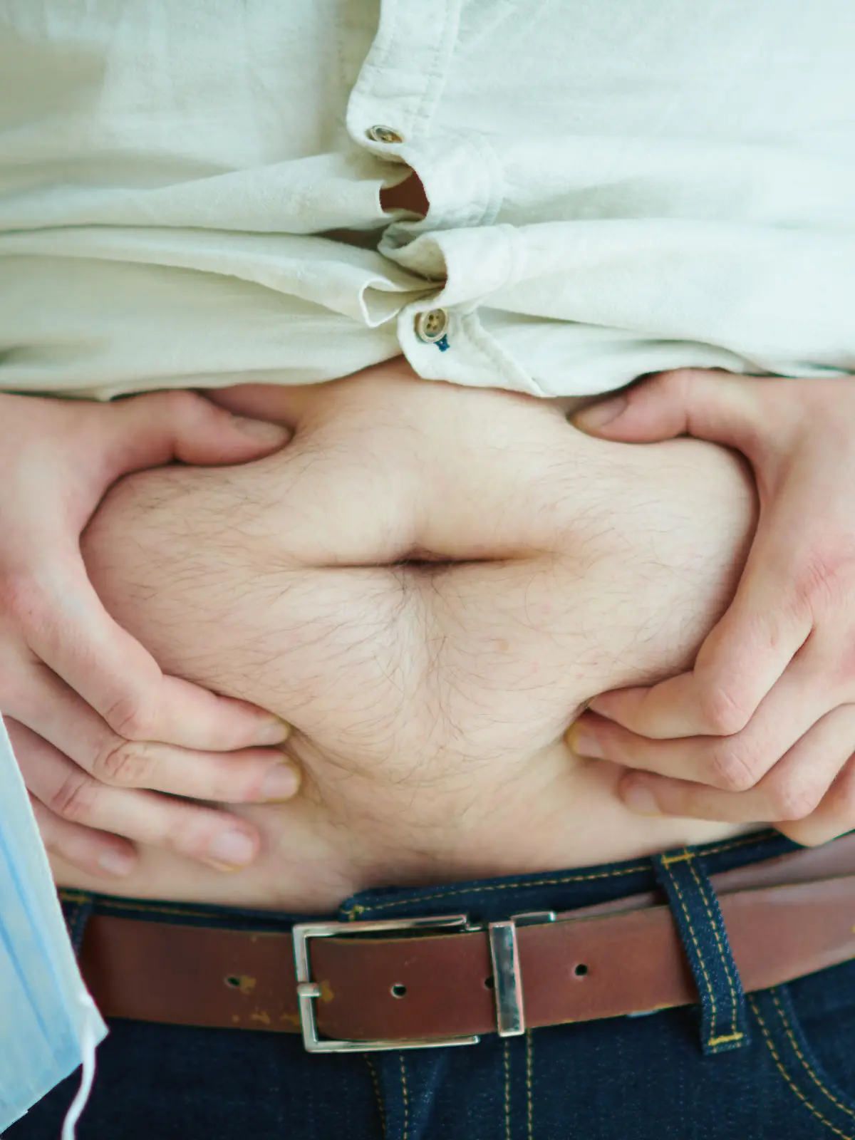 Ein Mann greift sich an seinen nicht mit Kleidung bedeckten Bauch, an einem Handgelenk taumelt eine Mund-Nasenschutz-Maske.