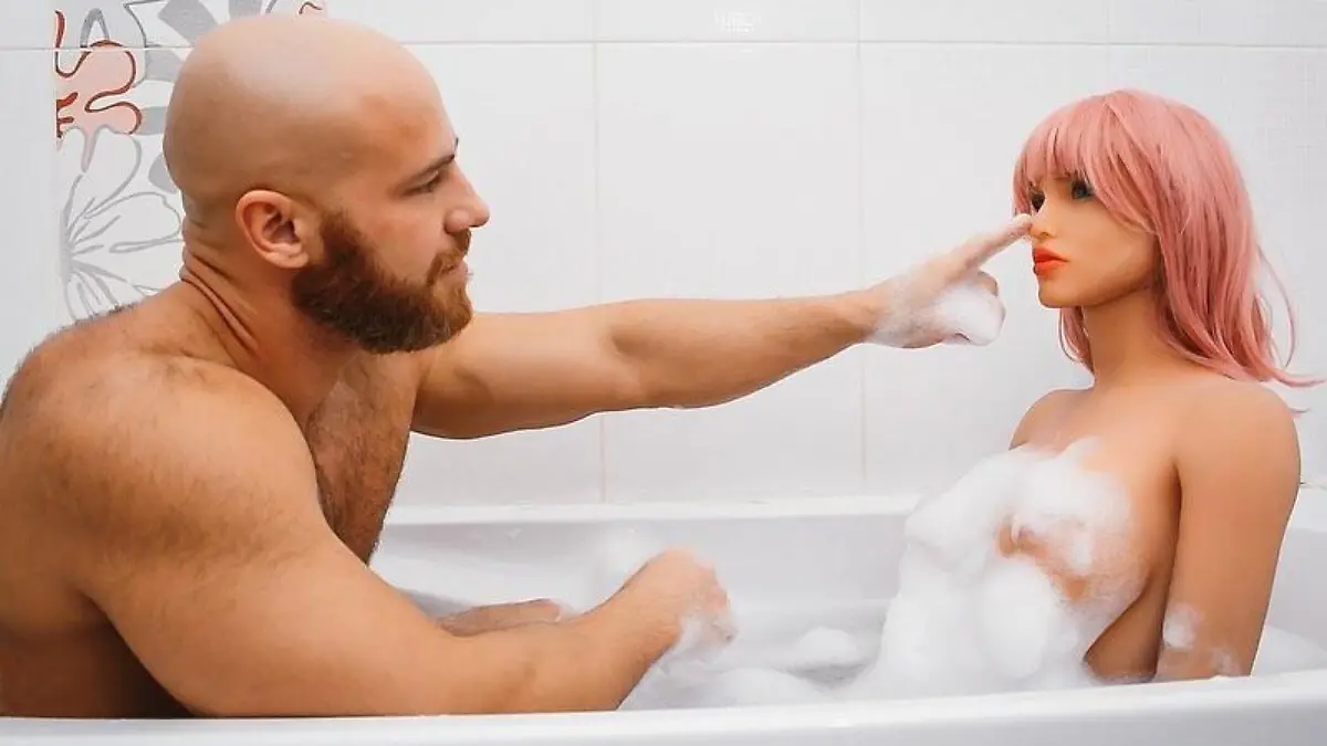 Mann sitzt mit einer Puppe in einer Badewanne.