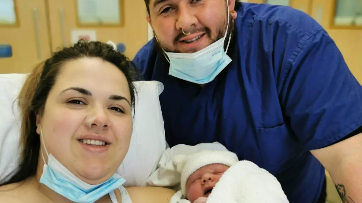 Mutter Amy hält ihren Sohn auf dem Arm während Vater Zac hinter ihr steht.