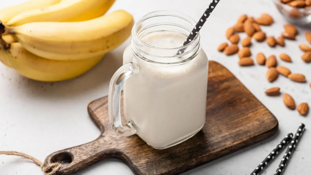 Ein leckerer Bananen-Protein-Smoothie ist hübsch angerichtet auf einem kleinen Holzbrettchen. Daneben liegen Strohalme, Bananen und Mandeln.