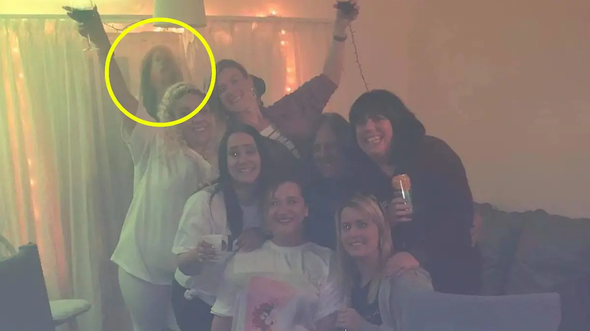 Man sieht sieben Frauen, die in die Kamera lächeln. Im Hintergrund ist eine schwummrige, dunkle Gestalt, die aussieht wie eine brünette Frau.