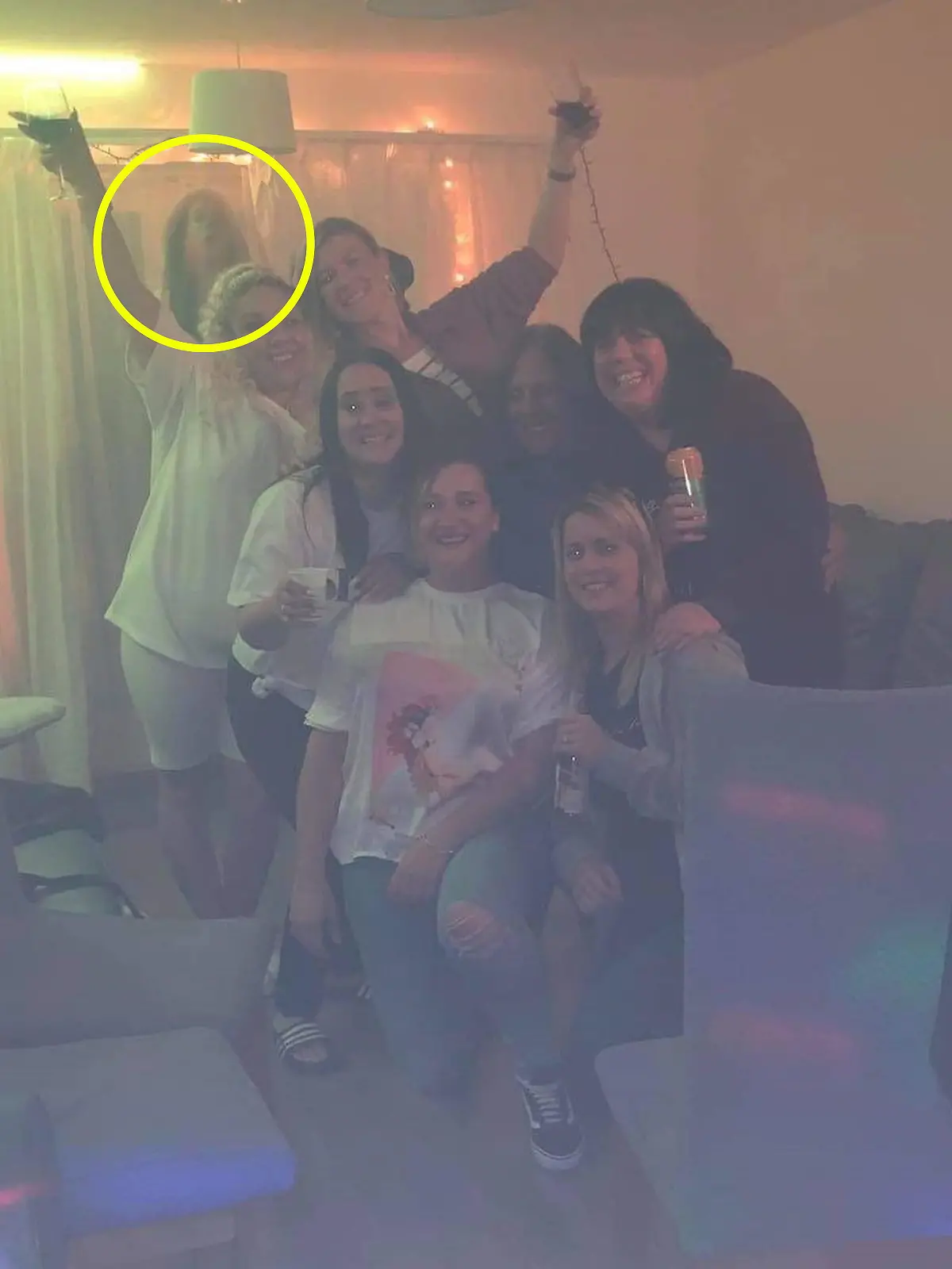 Man sieht sieben Frauen, die in die Kamera lächeln. Im Hintergrund ist eine schwummrige, dunkle Gestalt, die aussieht wie eine brünette Frau.