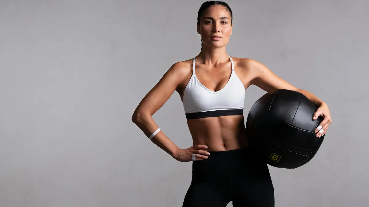 Portrait of beautiful mid adult woman looking at camera while holding heavy medicine ball isolated on grey background. Proud and fit woman standing on gray wall ready for gym exercise while looking at camera. Strong mixed race girl holding sports ball and relaxing after cross training workout with copy space.