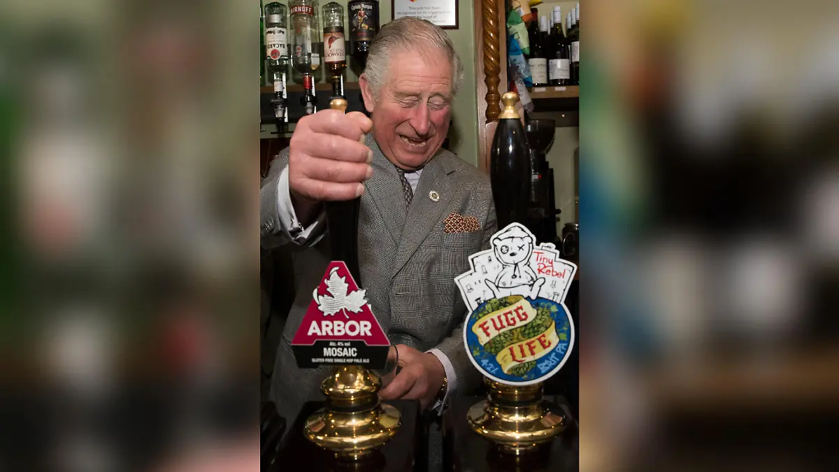 König Charles steht vor zwei Bier-Zapfhähnen und zapft breit grinsend ein Bier.