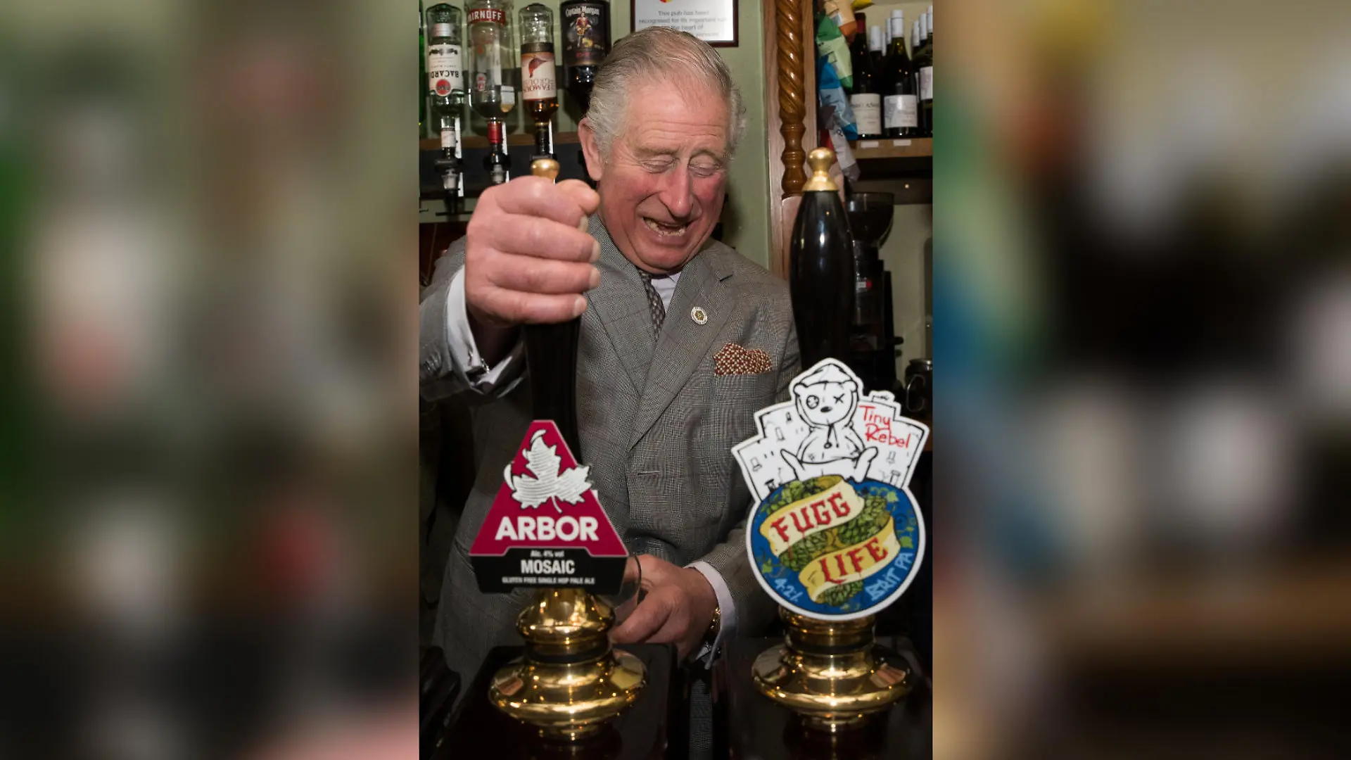 König Charles steht vor zwei Bier-Zapfhähnen und zapft breit grinsend ein Bier.