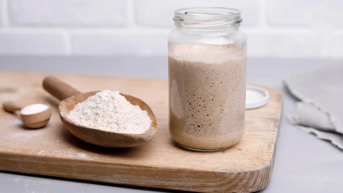 Auf einem Holzbrettchen liegen unter anderem ein großer Löffel mit Mehl, ein kleinerer mit Zucker und ein größeres Einmachglas, in dem bereits Teig drin liegt.
