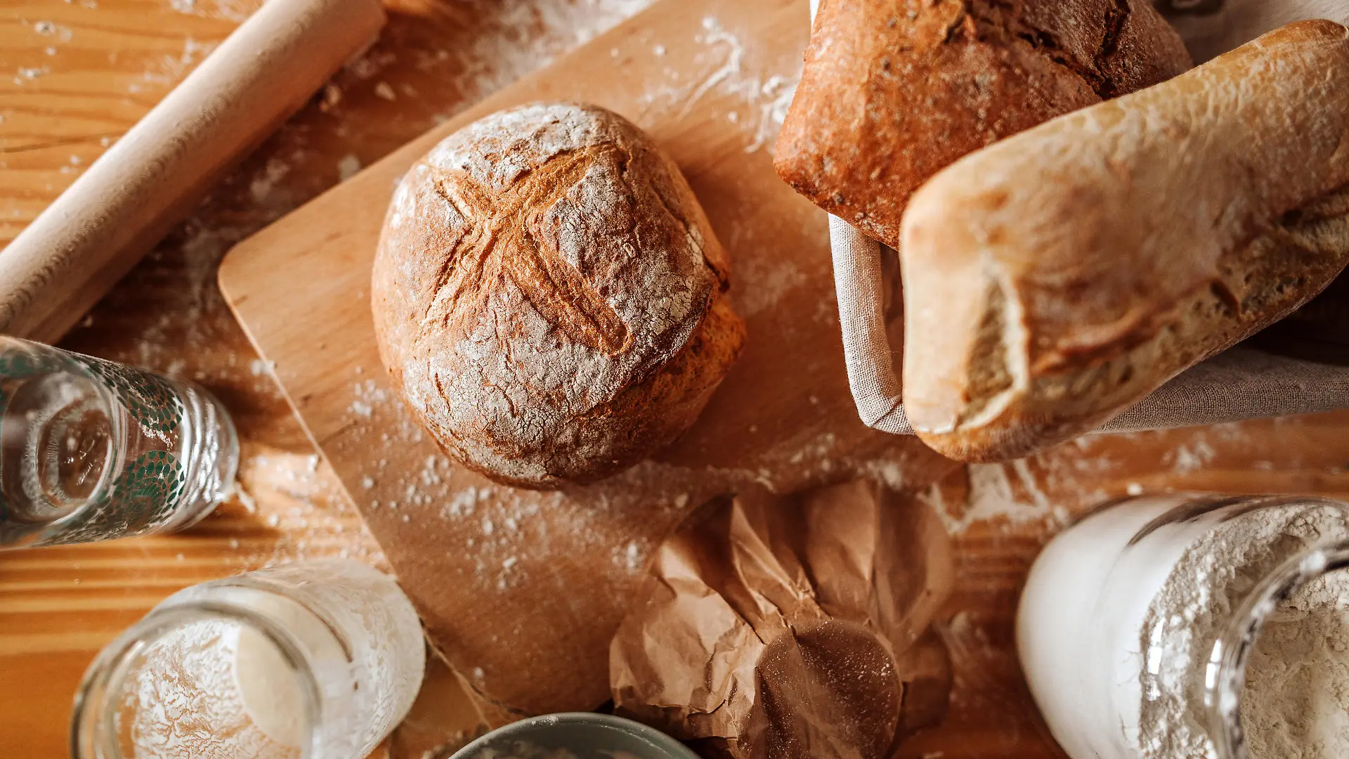 Auf einem Brettchen auf einem Esstisch liegen verschiedene Utensilien, unter anderem ein frisch gebackenes Sauerteigbrot.