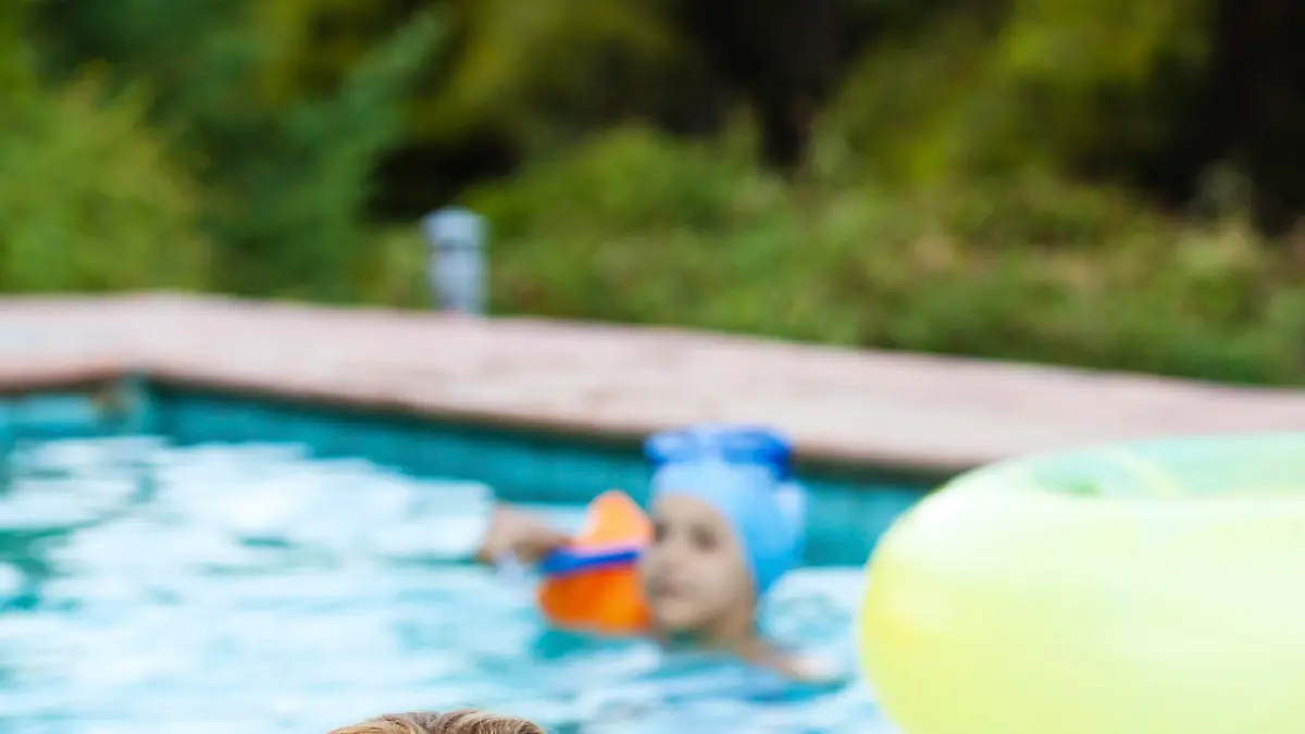 Little boy wearing water wings sitting by the swimming pool looking angry and sad, while mother talks to him.