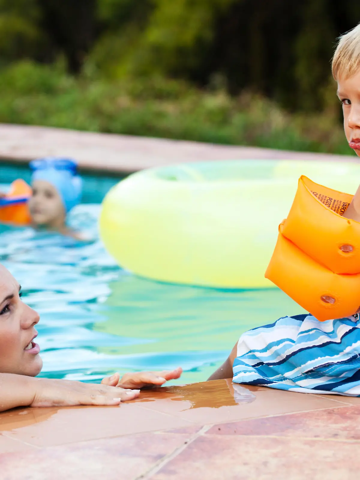 Little boy wearing water wings sitting by the swimming pool looking angry and sad, while mother talks to him.