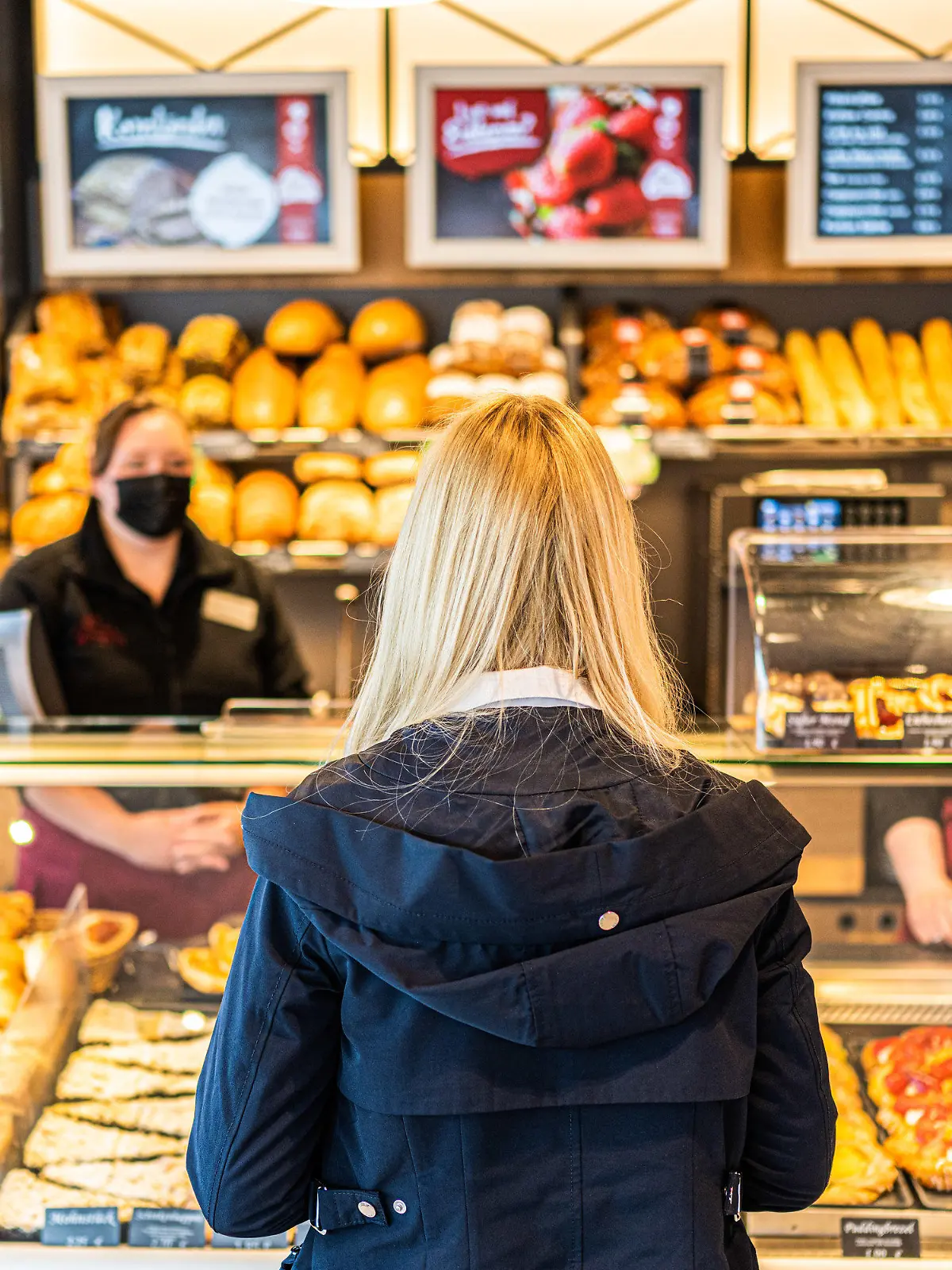 30.04.2021, Niedersachsen, Oldenburg: Josephine Marine (l) und Jana Kilian, beide Angestellte in der Oldenburger Bäckerei ·Musswessels·, stehen hinter der Verkaufstheke und bedienen eine Kundin. Mit neuen Ideen für altes Brot sollen weniger Lebensmittel im Müll landen. Rund 360 Bäckereifilialen in Niedersachsen informieren am Tag der Lebensmittelverschwendung an diesem Sonntag, wie Backwaren richtig gelagert und Reste schmackhaft verwertet werden können. Foto: Mohssen Assanimoghaddam/dpa +++ dpa-Bildfunk +++