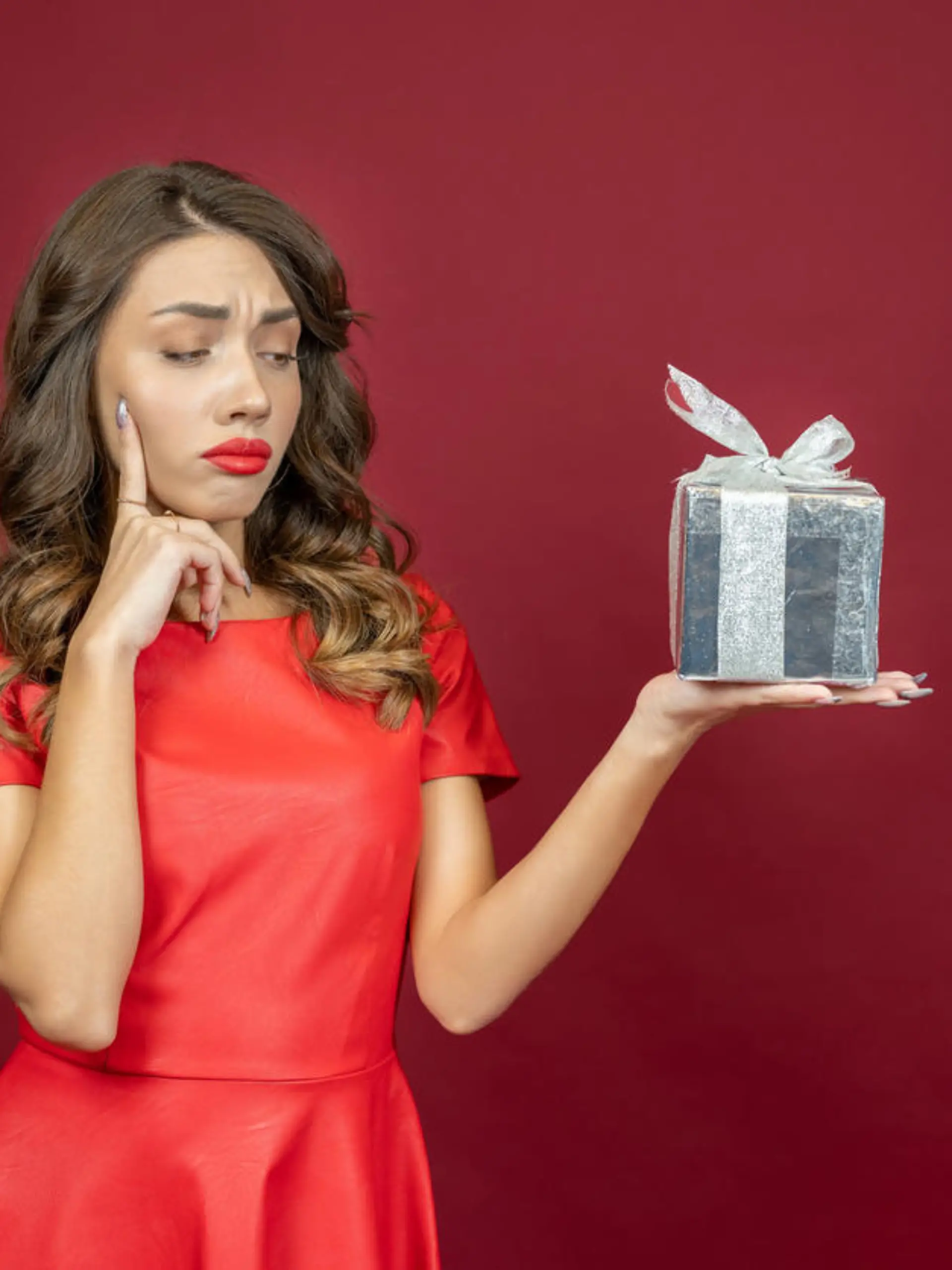 Annoyed brunette woman looking dismissively at a gift in red dress on a red background.