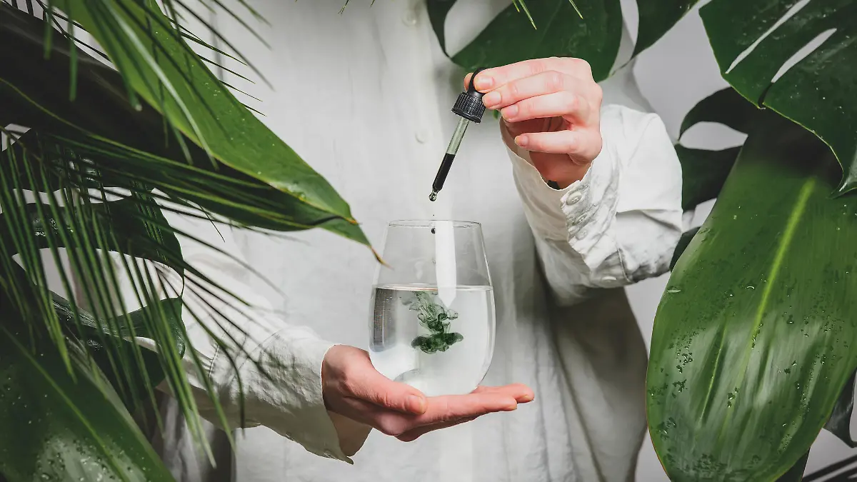 woman in a white shirt adds drops of chlorophyll to glass of water standing next to palm trees