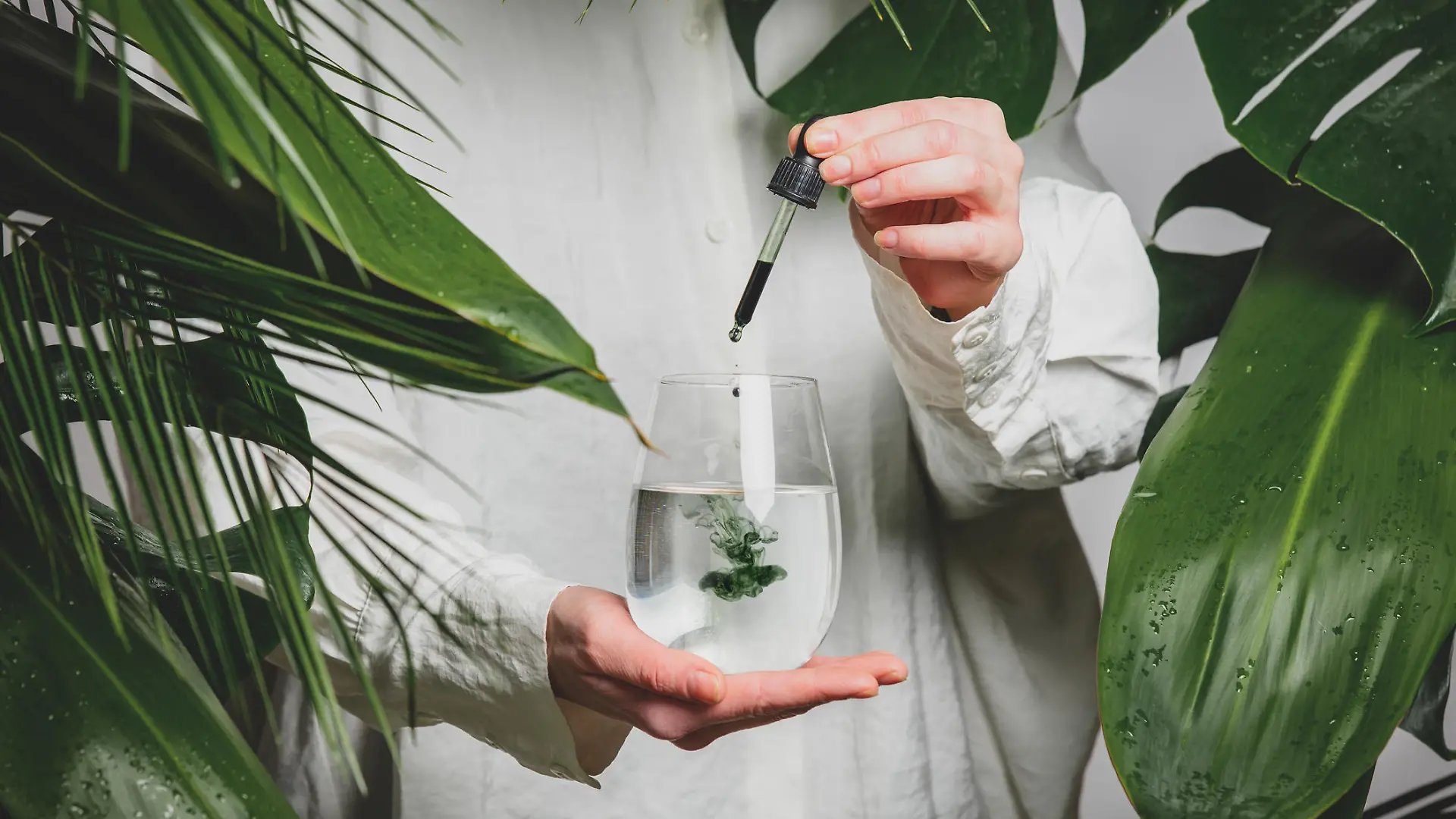 woman in a white shirt adds drops of chlorophyll to glass of water standing next to palm trees