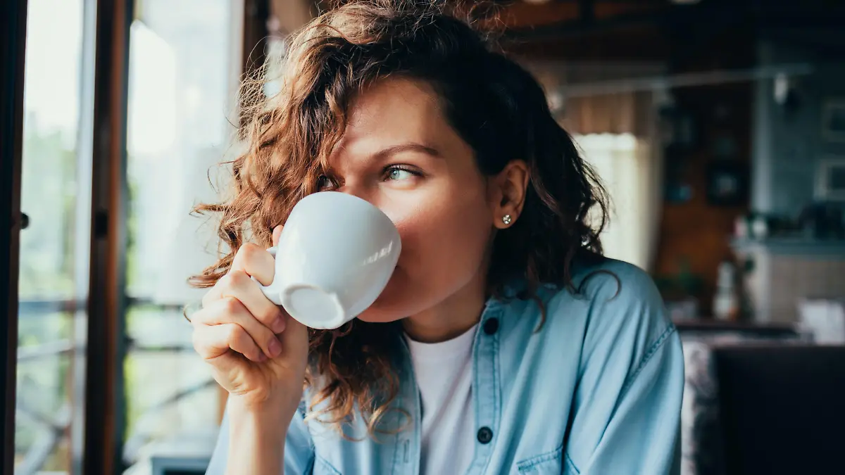 Portrait of happy beautiful young woman wearing blue shirt drinking coffee sitting at table in restaurant looking out the window on summer day.