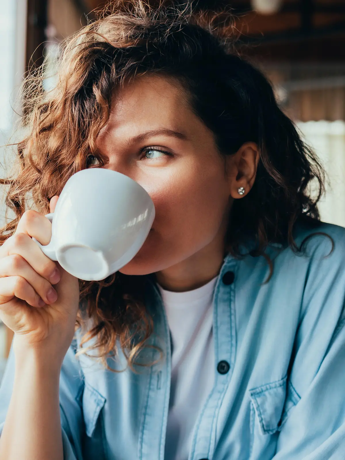 Portrait of happy beautiful young woman wearing blue shirt drinking coffee sitting at table in restaurant looking out the window on summer day.