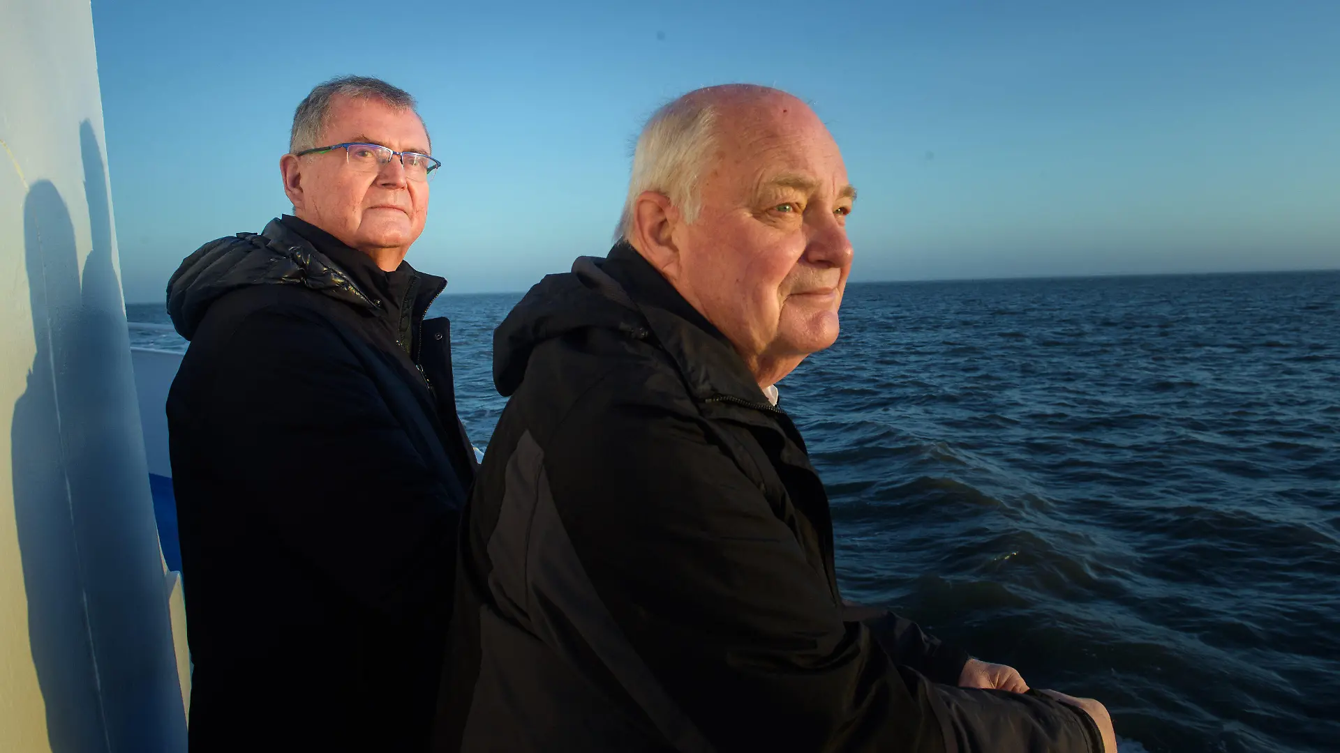 30.03.2021, Schleswig-Holstein, Hooge: Urs Philipzig (l), Mediziner aus Bredstedt und neuer Arzt von Hallig Hooge, und Gerhard Steinort (r), Mediziner in Langenhorn und ehemaliger Arzt der Hallig Hooge, stehen während des Sonnenaufgangs am Heck der MS Seeadler auf dem Weg zur Hallig Hooge. (zu dpa «Hooges Halligarzt geht in den Ruhestand») Foto: Gregor Fischer/dpa +++ dpa-Bildfunk +++