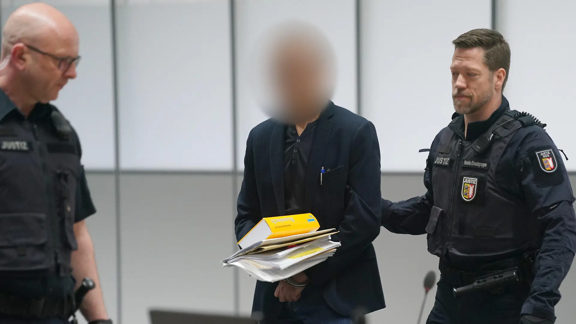 28.04.2023, Schleswig-Holstein, Itzehoe: Der Angeklagte (M) wird zu Beginn des Prozesses wegen Vergewaltigung und Mord von Justizbeamten in den Gerichtssaal des Landgerichts Itzehoe im China Logistic Center gebracht. Der Mann soll seine Ehefrau in Chemnitz vergewaltigt und einige Monate später in Heide ermordet haben. Die Ehepartner lebten in Trennung und hatten einen zur Tatzeit 13 Jahre alten gemeinsamen Sohn. Foto: Marcus Brandt/dpa - ACHTUNG: Person(en) wurde(n) aus rechtlichen Gründen gepixelt +++ dpa-Bildfunk +++