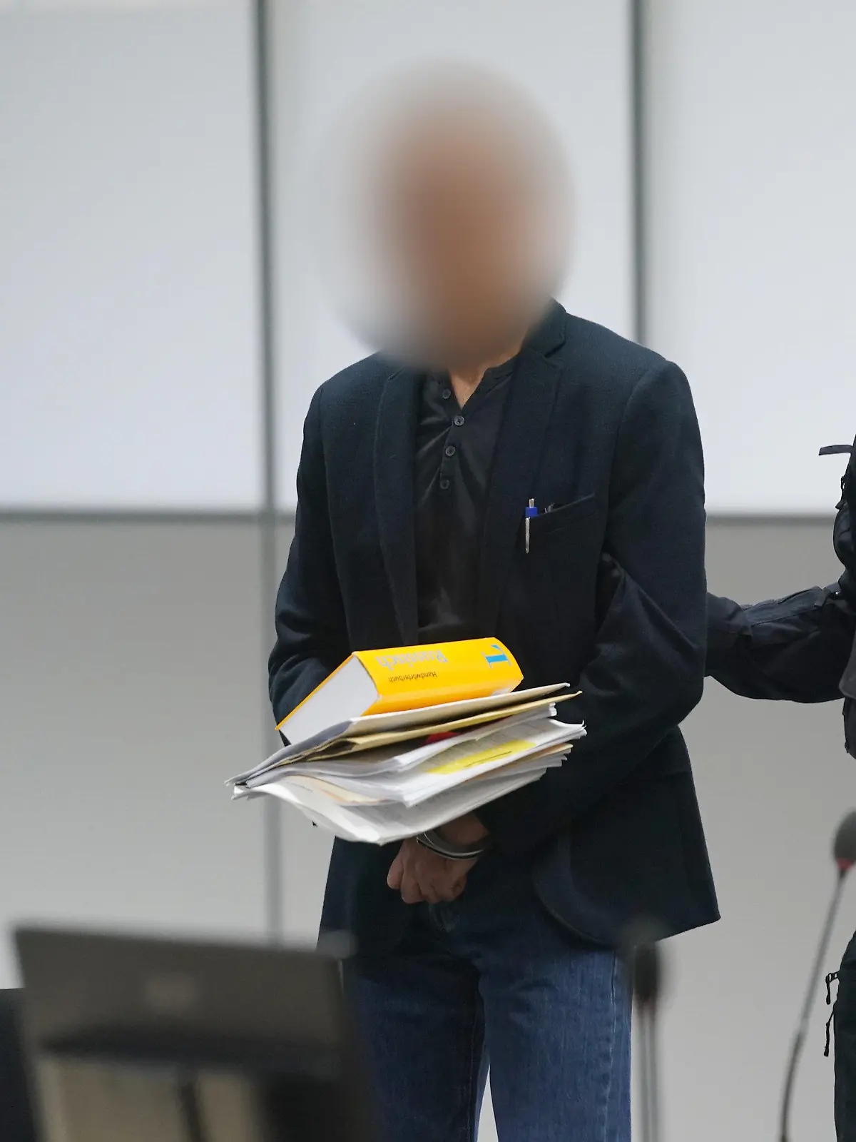 28.04.2023, Schleswig-Holstein, Itzehoe: Der Angeklagte (M) wird zu Beginn des Prozesses wegen Vergewaltigung und Mord von Justizbeamten in den Gerichtssaal des Landgerichts Itzehoe im China Logistic Center gebracht. Der Mann soll seine Ehefrau in Chemnitz vergewaltigt und einige Monate später in Heide ermordet haben. Die Ehepartner lebten in Trennung und hatten einen zur Tatzeit 13 Jahre alten gemeinsamen Sohn. Foto: Marcus Brandt/dpa - ACHTUNG: Person(en) wurde(n) aus rechtlichen Gründen gepixelt +++ dpa-Bildfunk +++