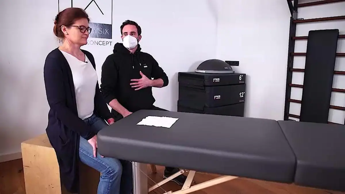 Frau neben Physiotherapeut an Liege. Taschentuch ausgebreitet darauf.