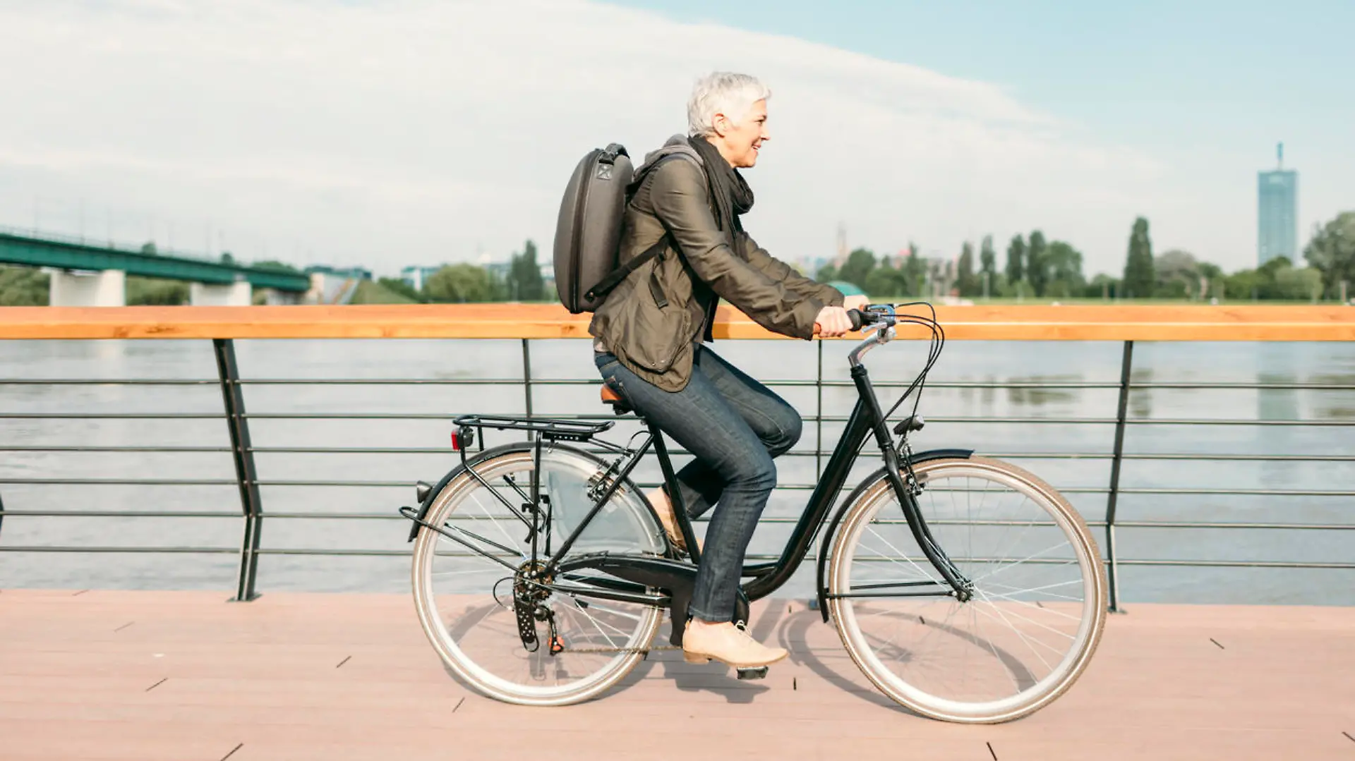 Mature businesswoman riding bicycle to and from her office. She is riding near river. Casual clothing with modern business backpack on her back. Riding city style bike.