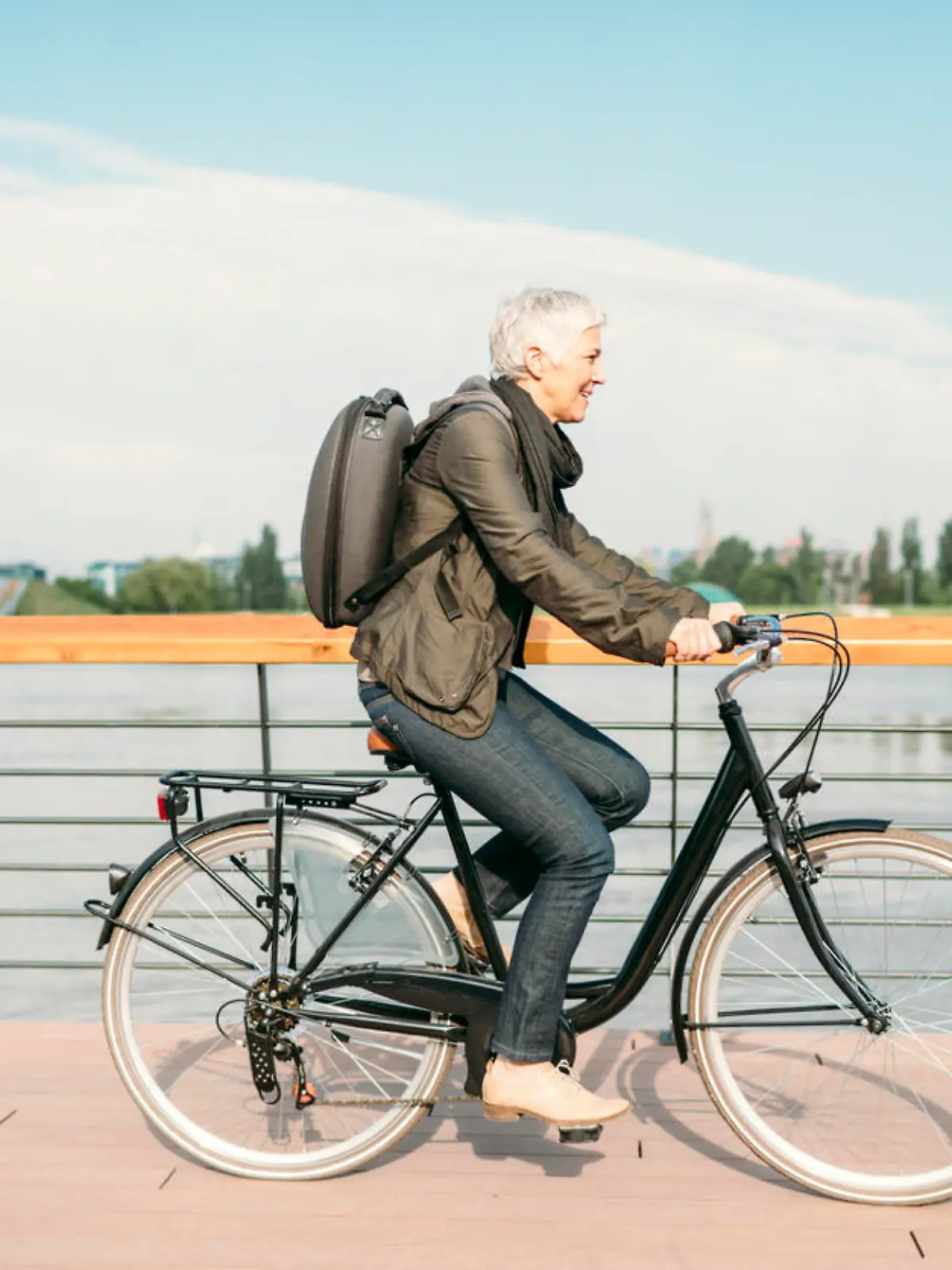 Mature businesswoman riding bicycle to and from her office. She is riding near river. Casual clothing with modern business backpack on her back. Riding city style bike.
