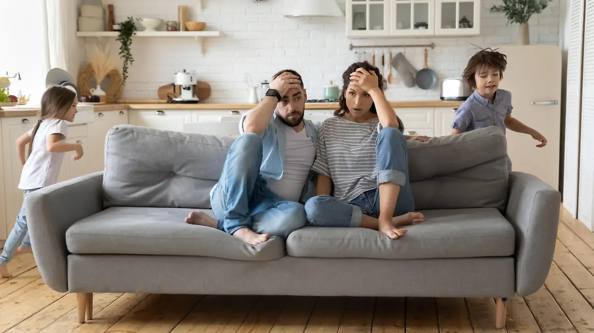 Tired mother and father sitting on couch feels annoyed exhausted while noisy little daughter and son shouting run around sofa where parents resting. Too active hyperactive kids, need repose concept