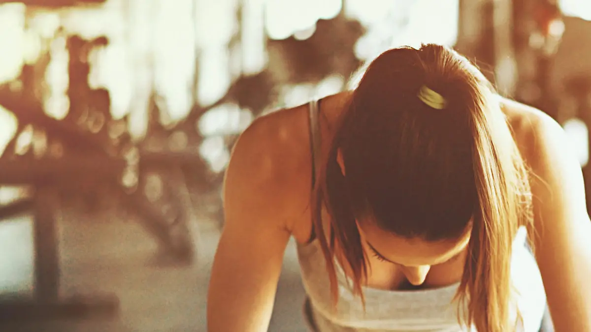 Closeup front view shot a slim muscular woman doing push ups in a gym. She's doing regular man style pushups with no excuses. Blurry gym equipment in background as well as black Pilates ball. Sunlight is flaring from the right hand side.