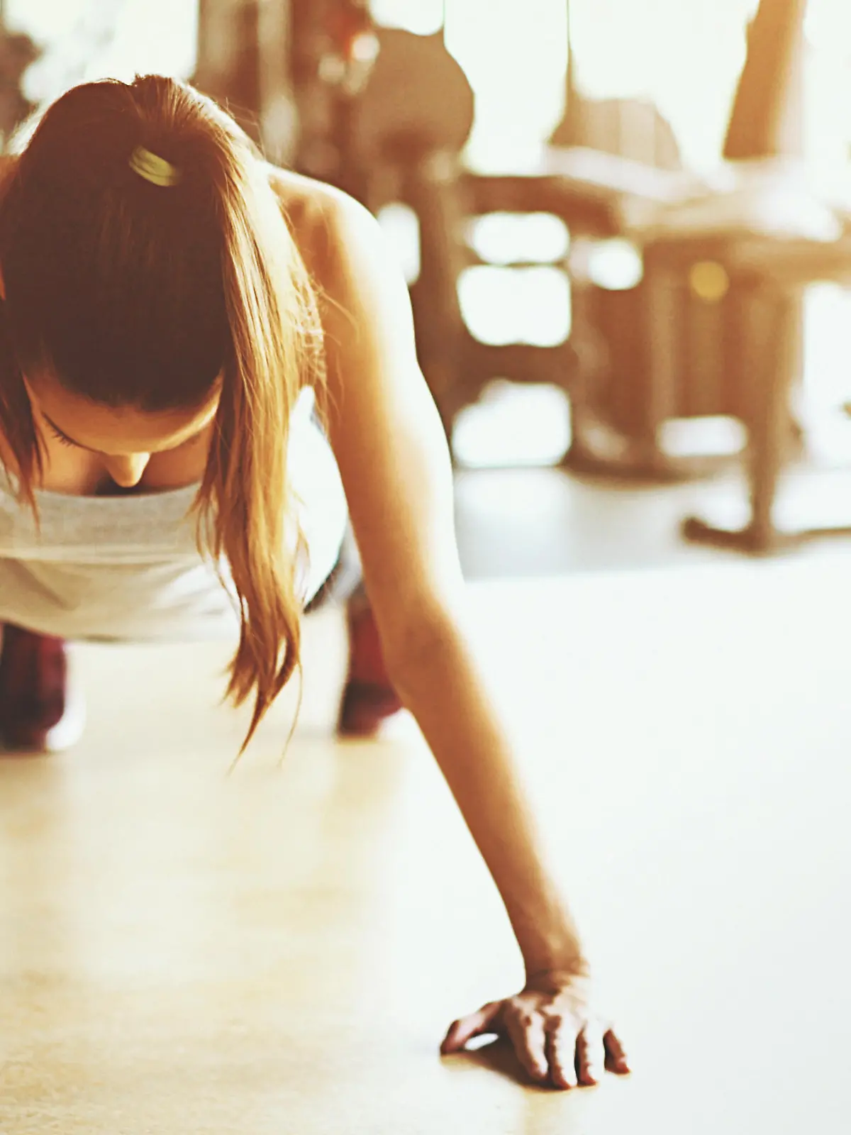 Closeup front view shot a slim muscular woman doing push ups in a gym. She's doing regular man style pushups with no excuses. Blurry gym equipment in background as well as black Pilates ball. Sunlight is flaring from the right hand side.