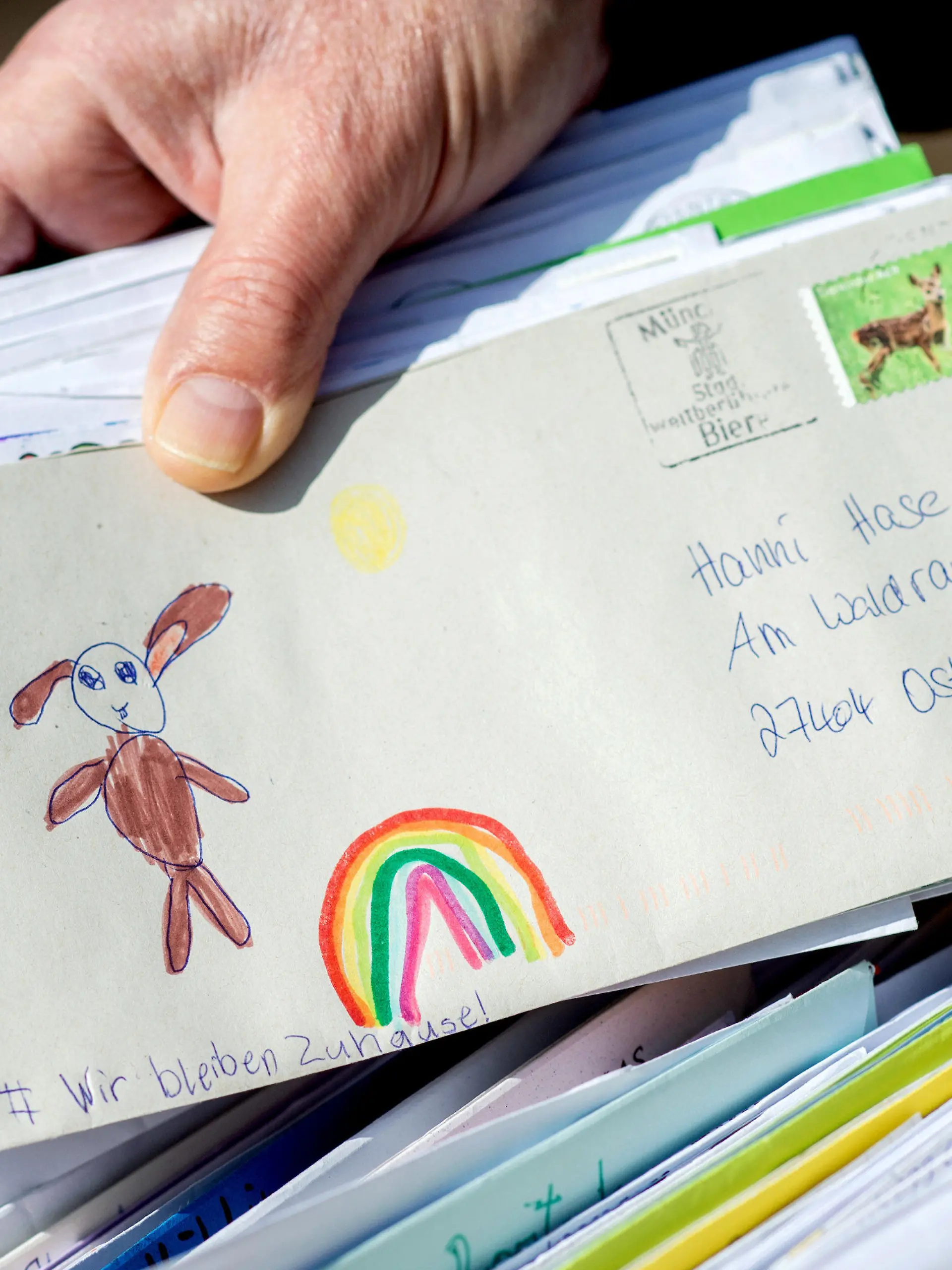 ARCHIV - 09.04.2020, Niedersachsen, Ostereistedt: Ein Mitarbeiter des Osterpostamts hält vor dem Gemeindehaus von Ostereistadt einen Brief in den Händen, der einen gemalten Regenbogen und die Aufschrift «Wir bleiben Zuhause!» zeigt. Die Briefschreibeaktion findet auch in diesem Jahr wieder statt bis zum 29. März. 2020 ging eine Rekordzahl von etwa 59000 Briefen im Osterpostamt ein. (Zu dpa: «Osterhase in Ostereistedt freut sich wieder auf Kinderbriefe») Foto: Hauke-Christian Dittrich/dpa +++ dpa-Bildfunk +++