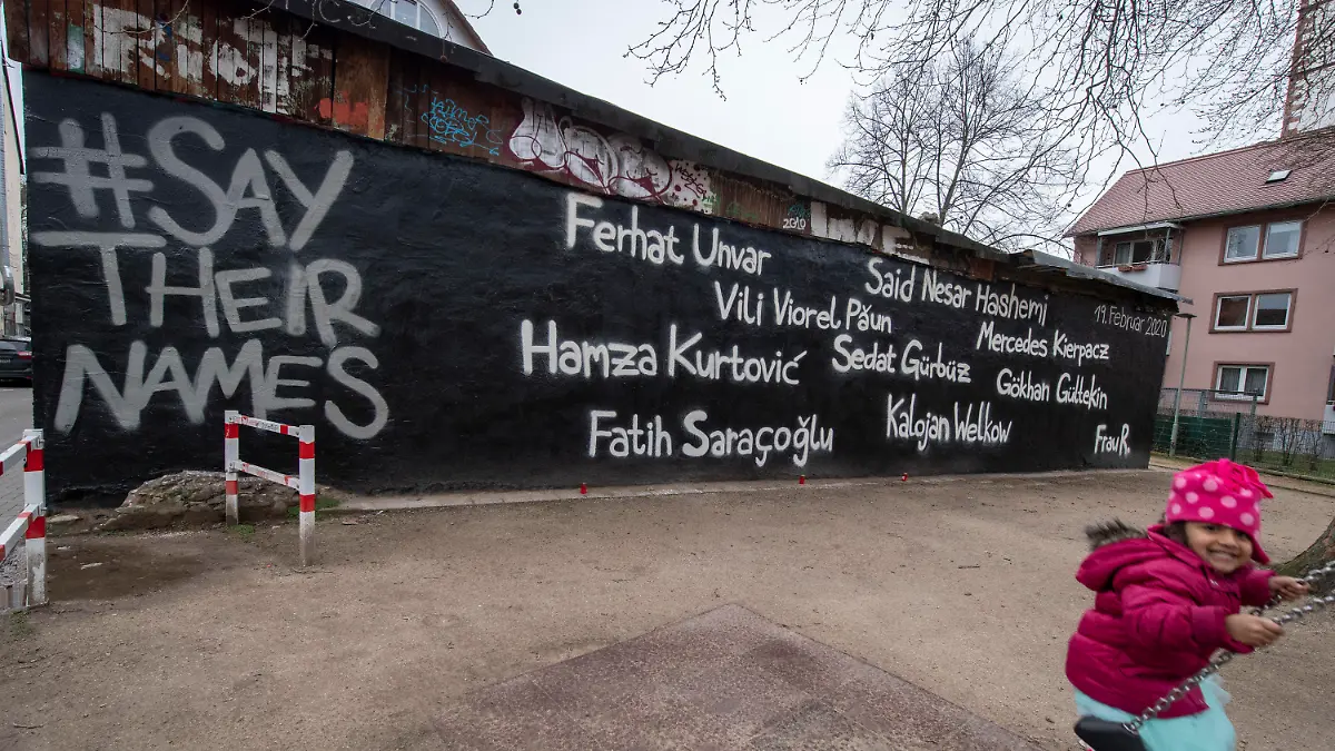 ARCHIV - 02.03.2020, Hessen, Hanau: Ein kleines Mädchen schaukelt vor der Wand eines Jugendzentrums, auf dem die Namen der Opfer des Anschlags vom 19. Februar geschrieben stehen. Links steht der Hashtag #Saytheirnames ("Nennt ihre Namen"), der Name der Mutter des Täters ist dabei mit "Frau R." abgekürzt. (zu dpa «Drei Monate nach Anschlag von Hanau») Foto: Boris Roessler/dpa +++ dpa-Bildfunk +++