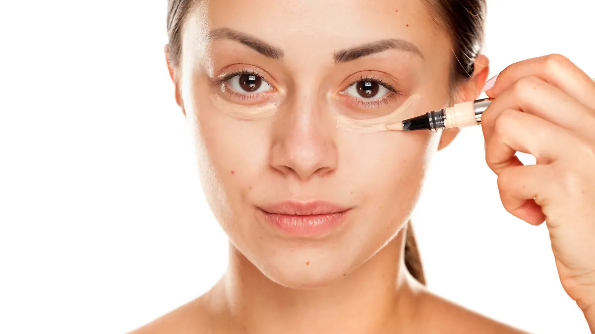 young beautiful girl applying concealer under her eyes on white background