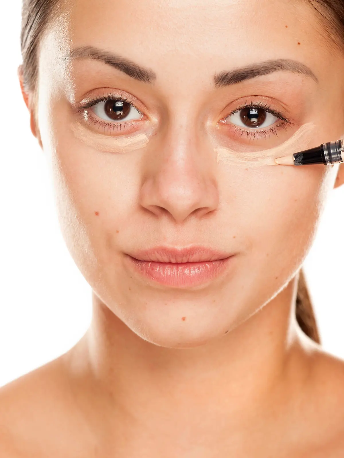 young beautiful girl applying concealer under her eyes on white background