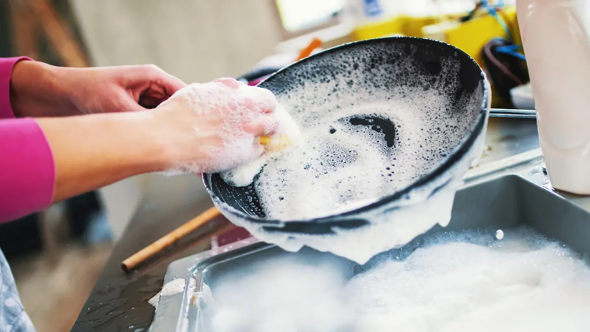 Closeup of unrecognizable woman doing dishes with lots of dirty plates and pots waiting aside. She's cleaning a black non stick cooking pan.