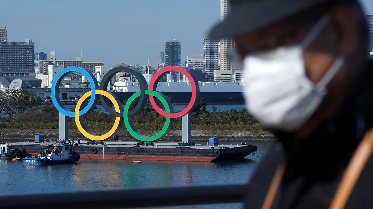 ARCHIV - 01.12.2020, Japan, Tokio: Ein Mann, der eine Gesichtsmaske trägt, um die Ausbreitung des Coronavirus einzudämmen, geht nahe der Olympischen Ringe in der Bucht von Tokio. Knapp ein halbes Jahr vor der geplanten Eröffnungsfeier halten sich die Zweifel an einer Austragung der Sommerspiele (zu dpa: «Olympia im Notstand: Was wird aus den Tokio-Spielen?») Foto: Eugene Hoshiko/AP/dpa +++ dpa-Bildfunk +++