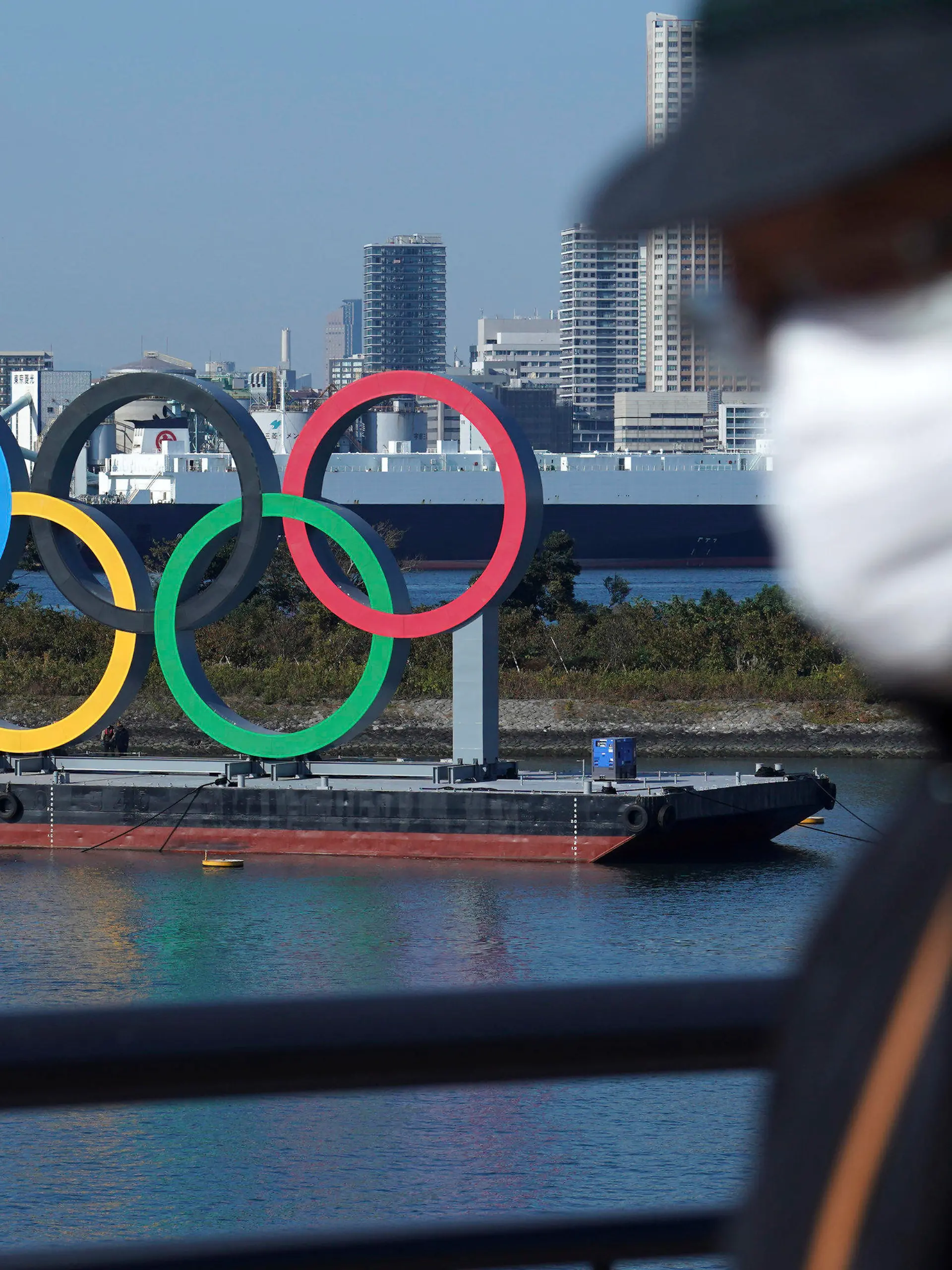 ARCHIV - 01.12.2020, Japan, Tokio: Ein Mann, der eine Gesichtsmaske trägt, um die Ausbreitung des Coronavirus einzudämmen, geht nahe der Olympischen Ringe in der Bucht von Tokio. Knapp ein halbes Jahr vor der geplanten Eröffnungsfeier halten sich die Zweifel an einer Austragung der Sommerspiele (zu dpa: «Olympia im Notstand: Was wird aus den Tokio-Spielen?») Foto: Eugene Hoshiko/AP/dpa +++ dpa-Bildfunk +++
