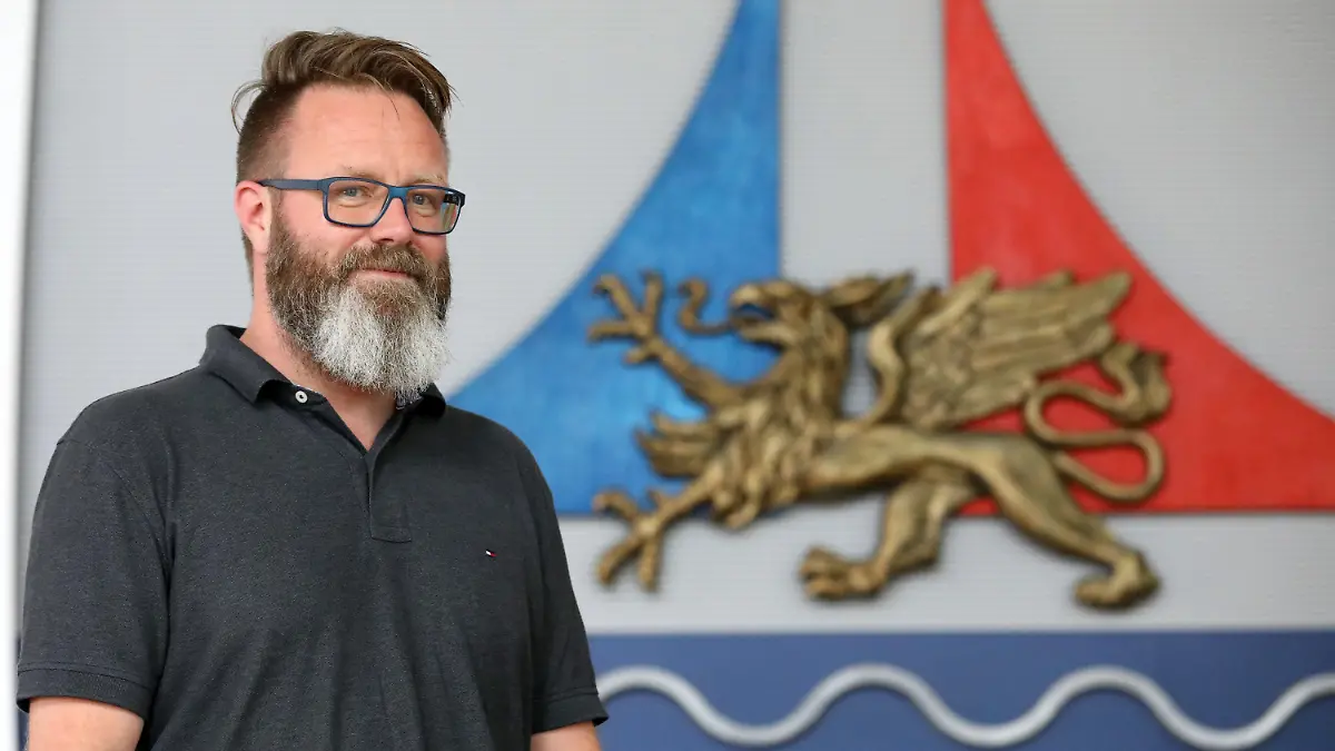 21.08.2020, Mecklenburg-Vorpommern, Rostock: Claus Ruhe Madsen, Oberbürgermeister, steht im Bürgerschaftssaal neben dem Stadtlogo. Der Däne wurde vor einem Jahr Oberbürgermeister der größten Stadt Mecklenburg-Vorpommerns. Foto: Bernd Wüstneck/dpa-Zentralbild/dpa +++ dpa-Bildfunk +++