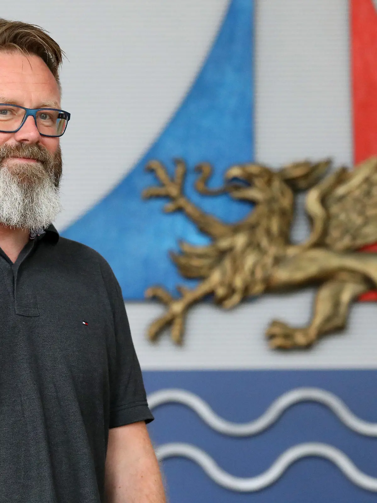 21.08.2020, Mecklenburg-Vorpommern, Rostock: Claus Ruhe Madsen, Oberbürgermeister, steht im Bürgerschaftssaal neben dem Stadtlogo. Der Däne wurde vor einem Jahr Oberbürgermeister der größten Stadt Mecklenburg-Vorpommerns. Foto: Bernd Wüstneck/dpa-Zentralbild/dpa +++ dpa-Bildfunk +++