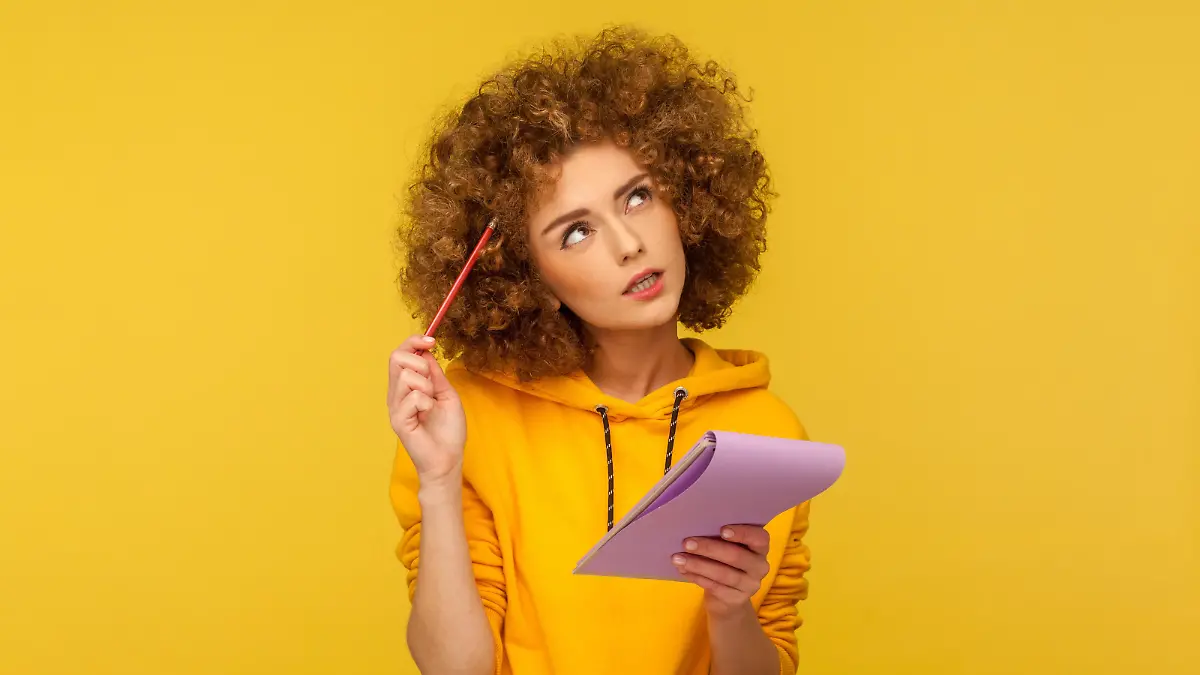 Beautiful pensive woman with Afro hairstyle holding notepad and pencil in hands thinking over plans for startup, wearing casual style hoodie. Indoor studio shot isolated on yellow background. || Modellfreigabe vorhanden