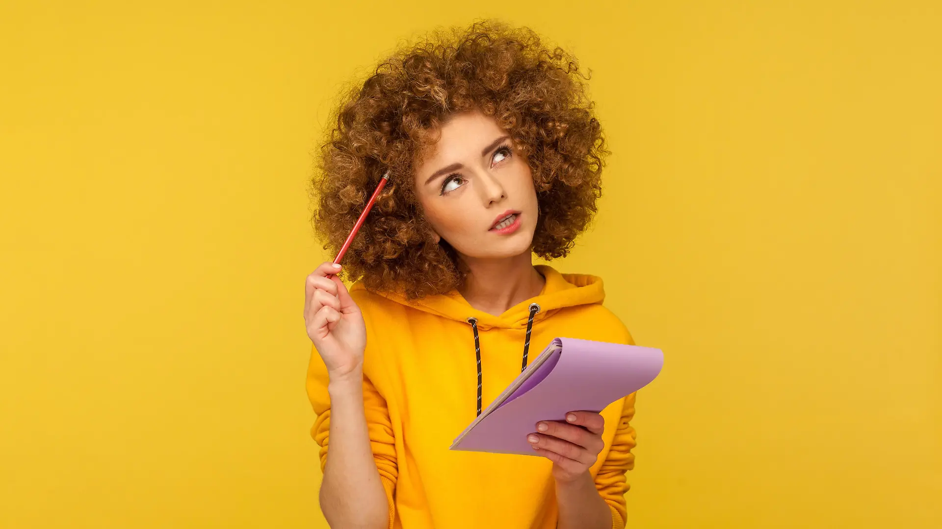 Beautiful pensive woman with Afro hairstyle holding notepad and pencil in hands thinking over plans for startup, wearing casual style hoodie. Indoor studio shot isolated on yellow background. || Modellfreigabe vorhanden