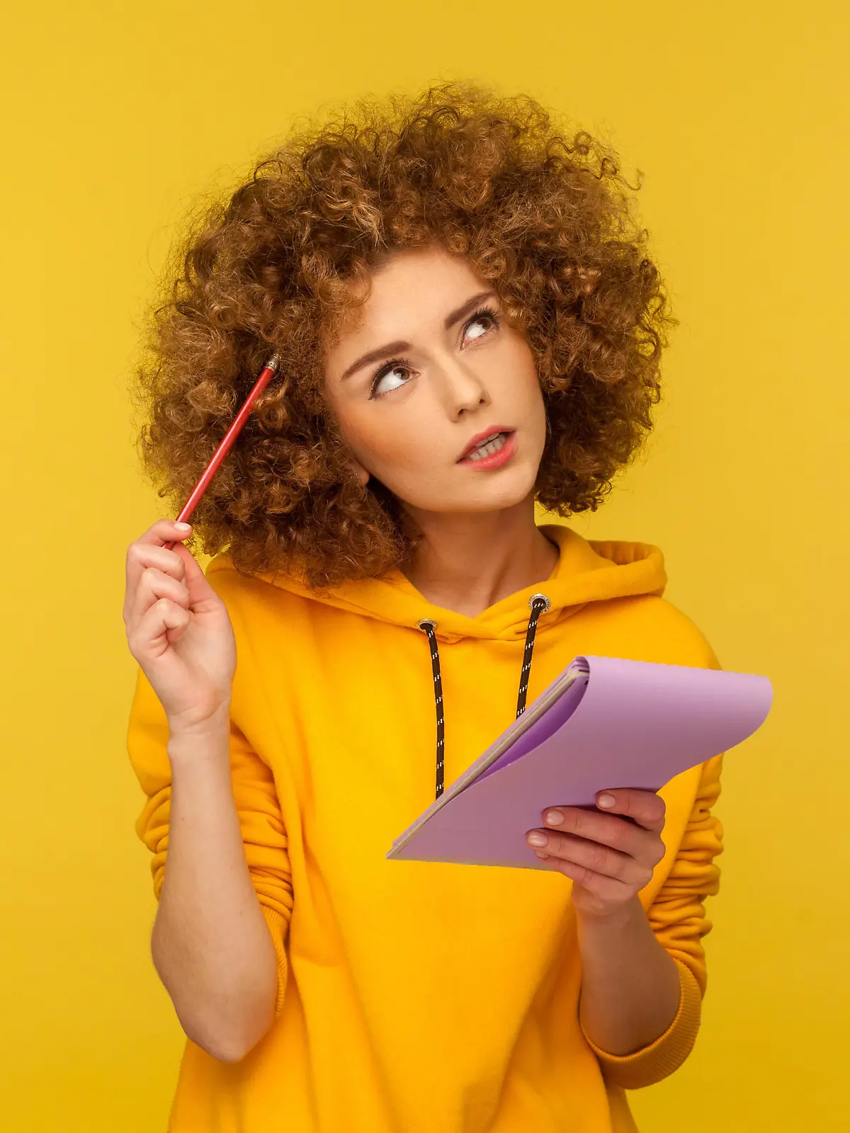 Beautiful pensive woman with Afro hairstyle holding notepad and pencil in hands thinking over plans for startup, wearing casual style hoodie. Indoor studio shot isolated on yellow background. || Modellfreigabe vorhanden