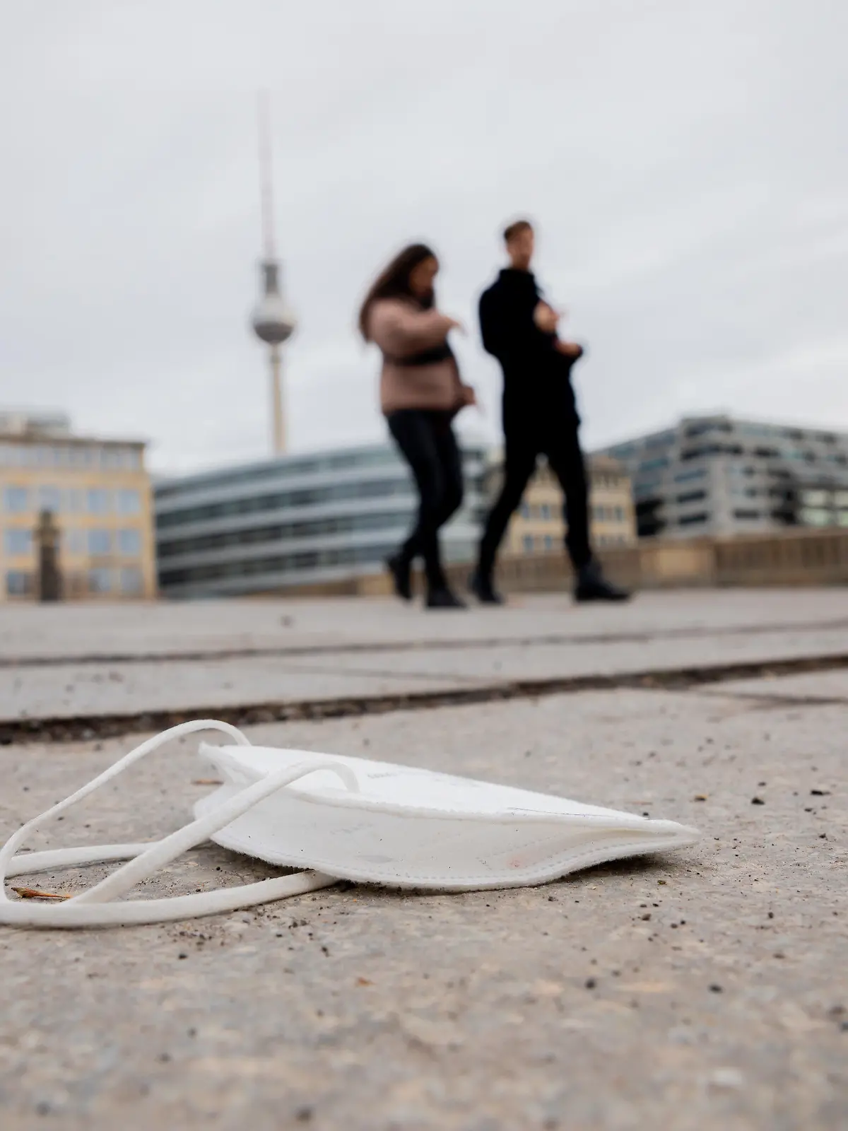 20.01.2021, Berlin: Eine FFP2-Maske liegt auf der Friedrichsbrücke, während im Hintergrund Passanten vorbeigehen. Foto: Christoph Soeder/dpa +++ dpa-Bildfunk +++