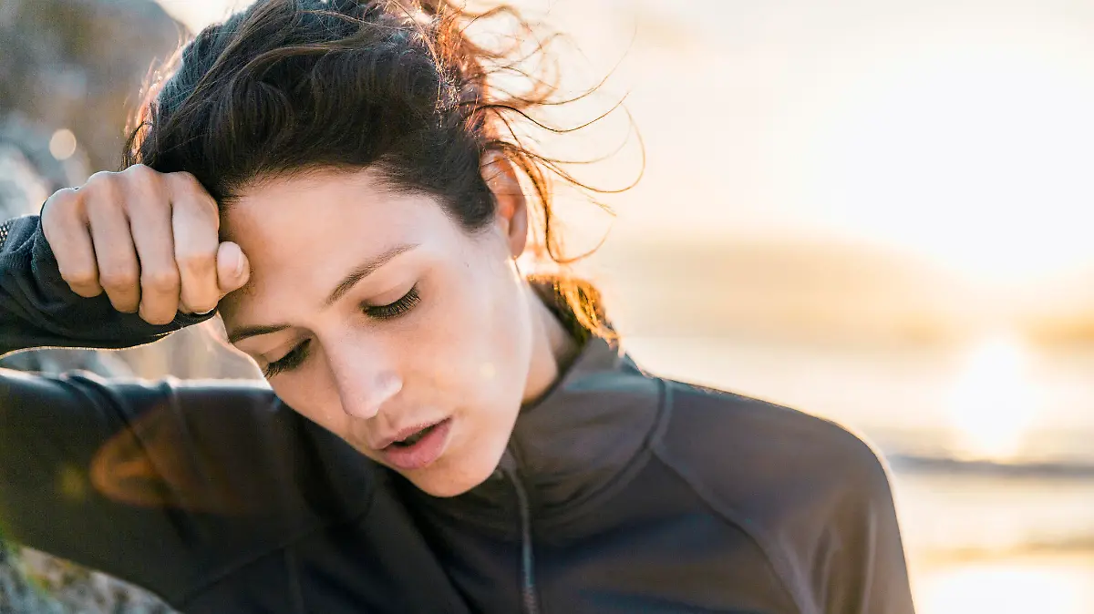 A photo of tired sporty woman looking down. Exhausted female athlete is wiping her forehead. She is wearing sportswear at beach.