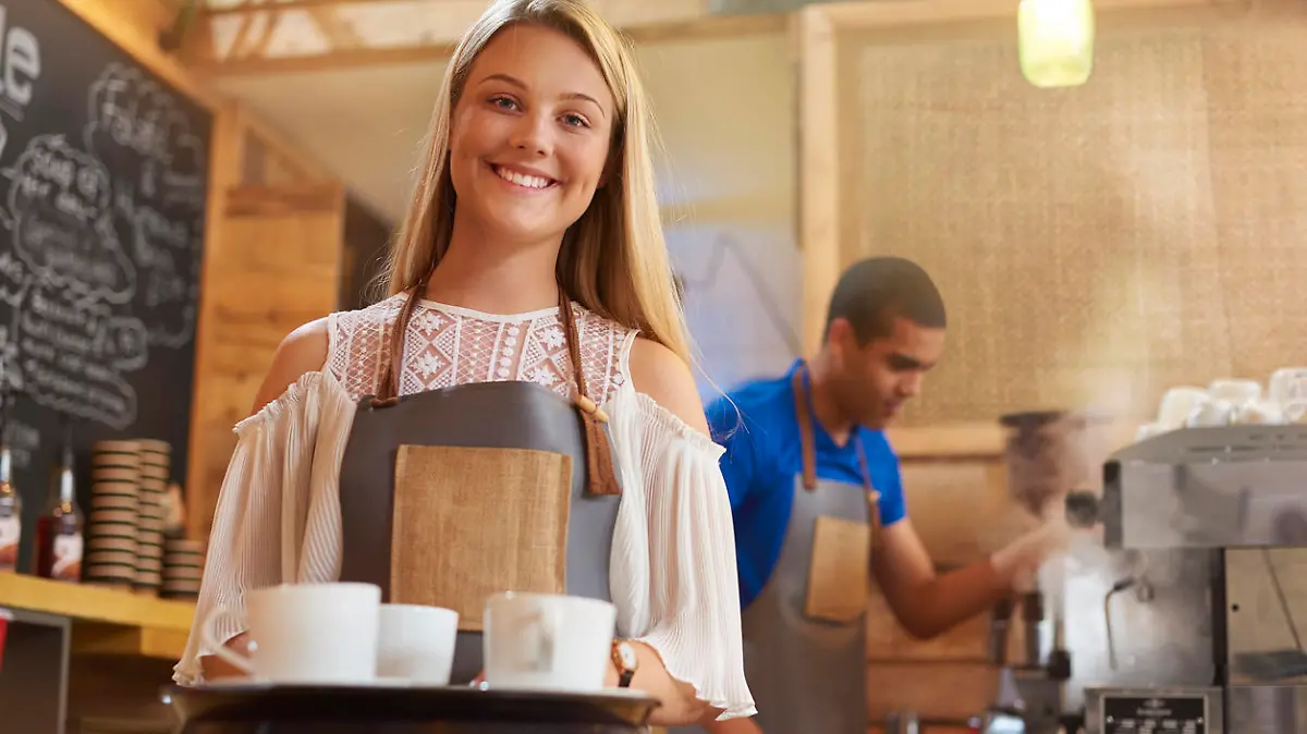 a coffee shop worker smiles to camera as she prepares to take an order to a table in a trendy little coffee outlet . In the background a male colleague is cleaning the coffee machine .