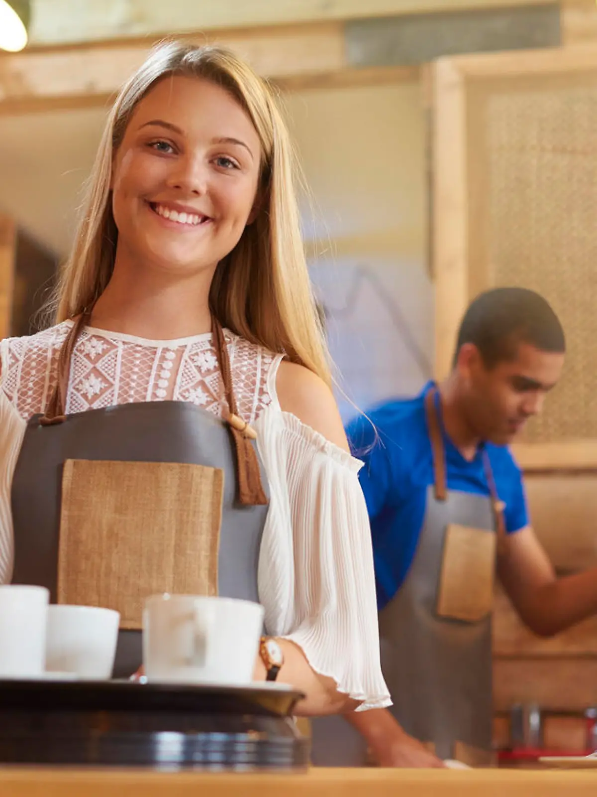 a coffee shop worker smiles to camera as she prepares to take an order to a table in a trendy little coffee outlet . In the background a male colleague is cleaning the coffee machine .