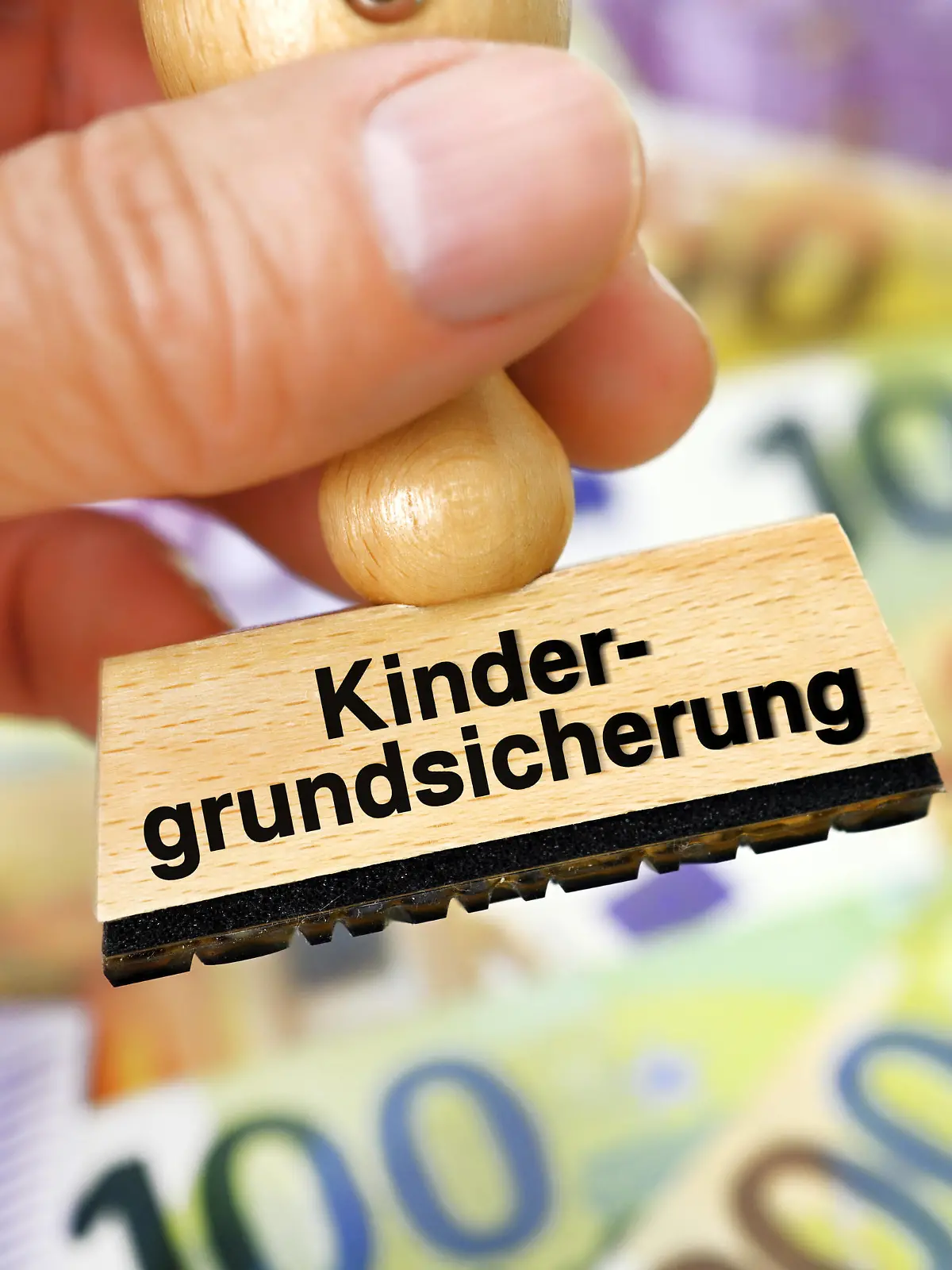FOTOMONTAGE, Hand hält Stempel mit Schriftzug Kindergrundsicherung über Geldscheinen *** PHOTOMONTAGE, hand holding stamp with writing child basic security over banknotes