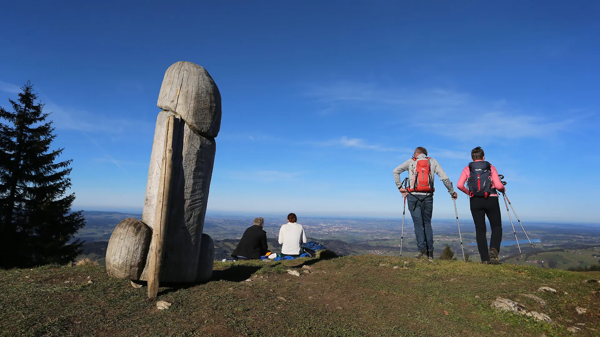 14.11.2020, Bayern, Rettenberg: Wanderer genießen neben der zwei Meter hohen Penis-Skulptur aus Holz auf dem Grat des Grünten den Ausblick. Seit vier Jahren steht das ungewöhnliche «Kulturdenkmal» auf dem Berg. (zu dpa «Kulturdenkmal»: Holzpenis auf Allgäuer Berg steht wieder) Foto: Karl-Josef Hildenbrand/dpa +++ dpa-Bildfunk +++
