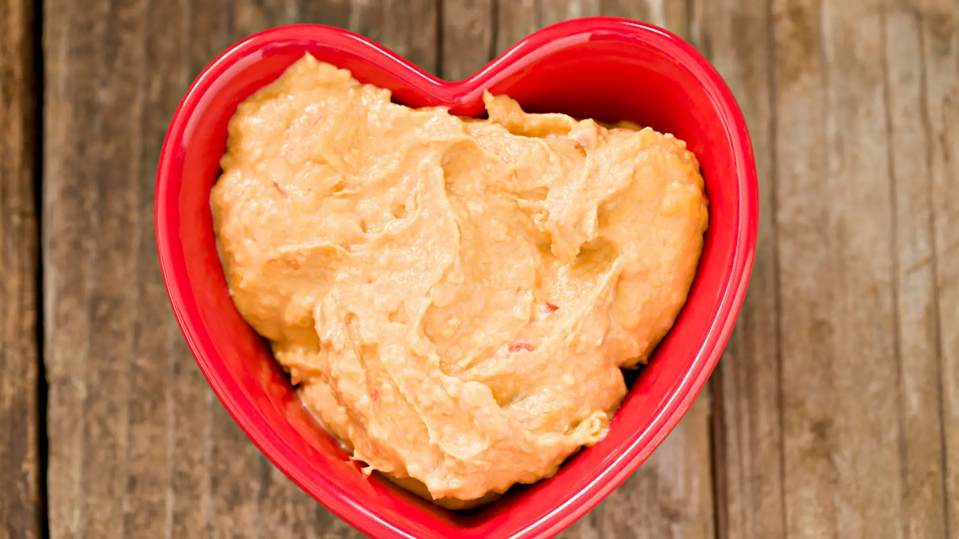 An overhead shot of a red heart shaped bowl with heart healthy rosasted red pepper humus. Shot on a grungy old wooden table. Humus is a Mediterranean spread made out of chickpeas or garbanzo beans.